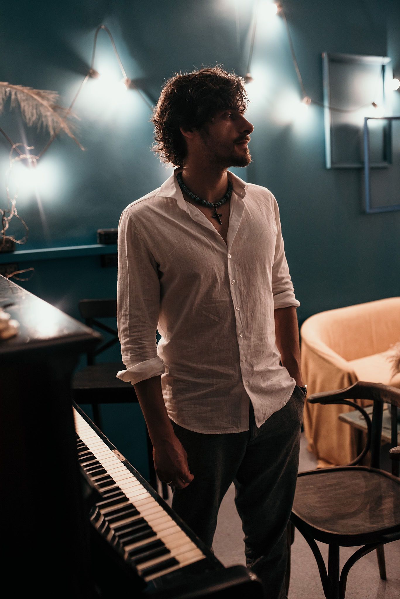 Artistic portrait in moody lighting showing figure near piano in white shirt against dark teal walls.