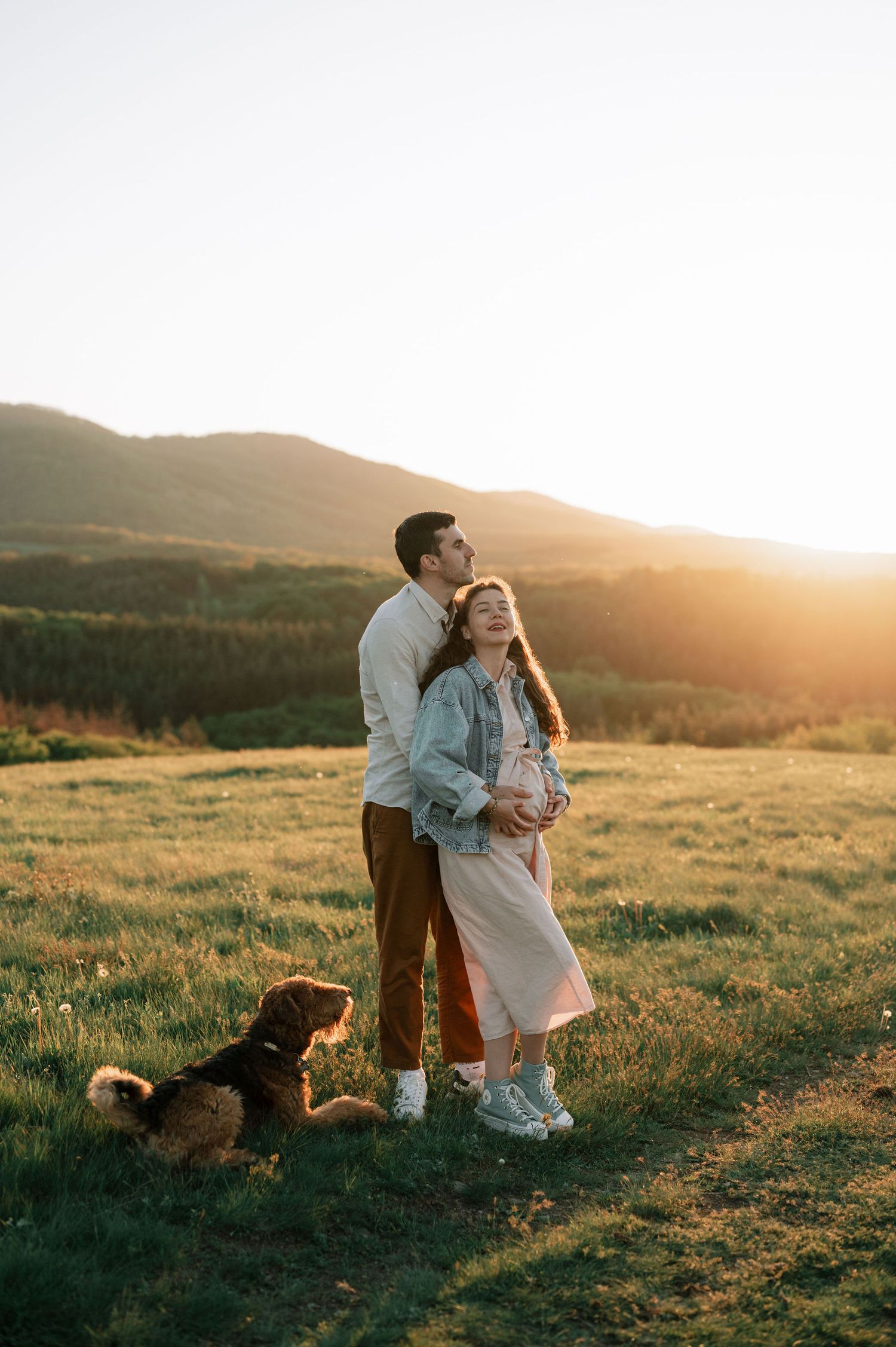 A couple and their dog share a tender moment at sunset in a grassy hilltop meadow.