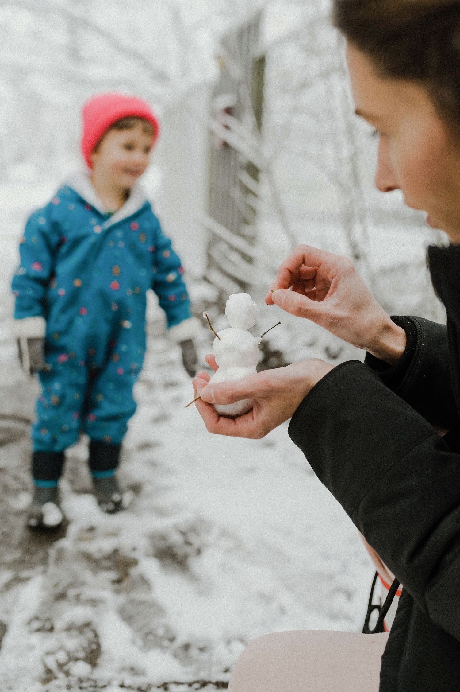 A child in a blue snowsuit and pink hat watches someone making a snowball on a winter day.