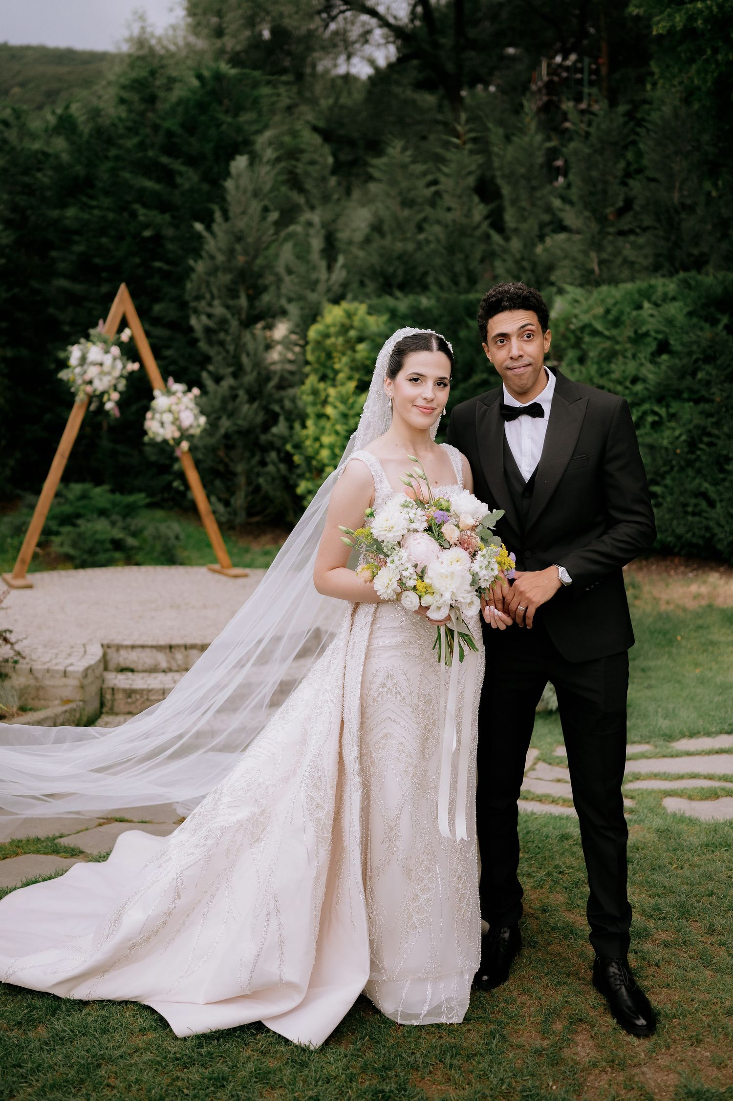 Elegant wedding couple poses in front of wooden triangle arch with flowing white dress and bouquet.