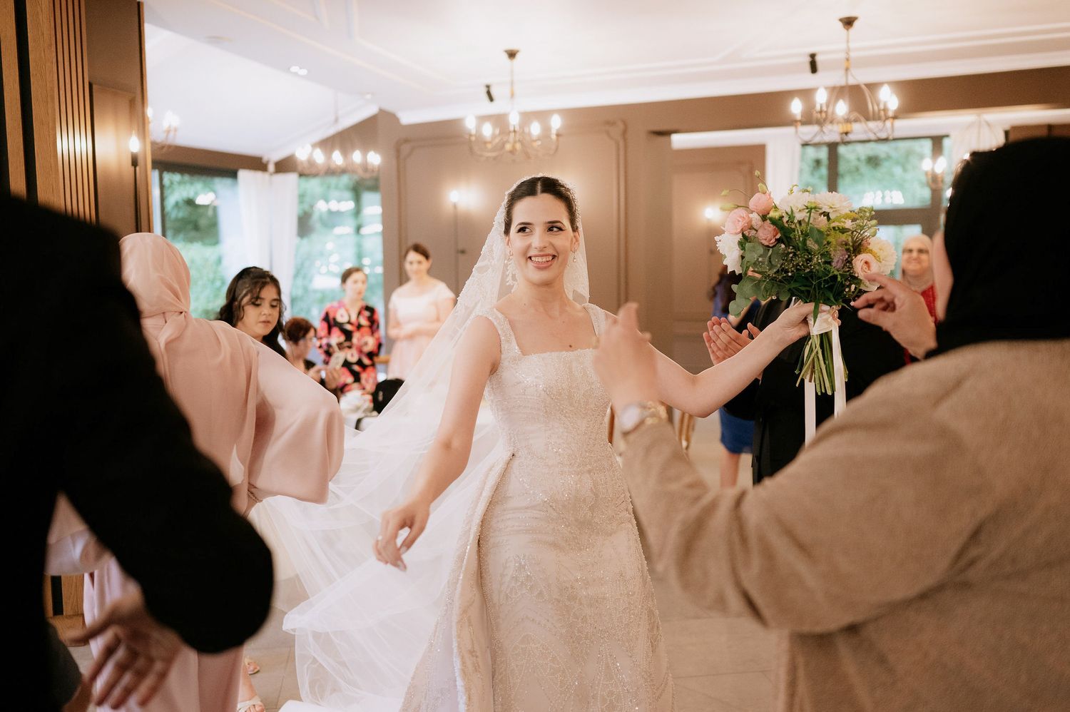 Wedding party celebrates in an elegant indoor venue with chandelier lighting and warm brown decor.