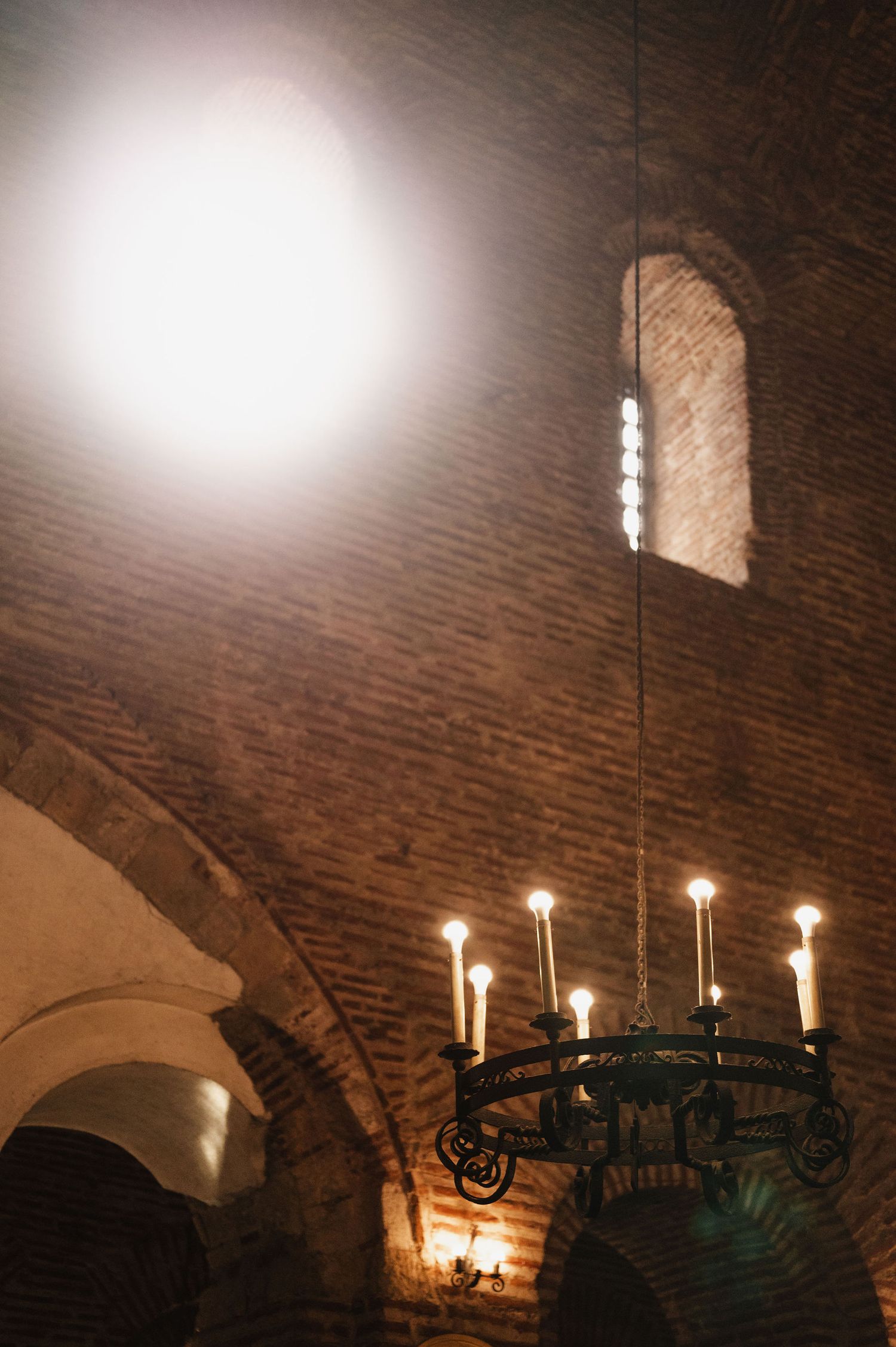 Wrought iron chandelier with glowing candles illuminates a rustic brick ceiling.