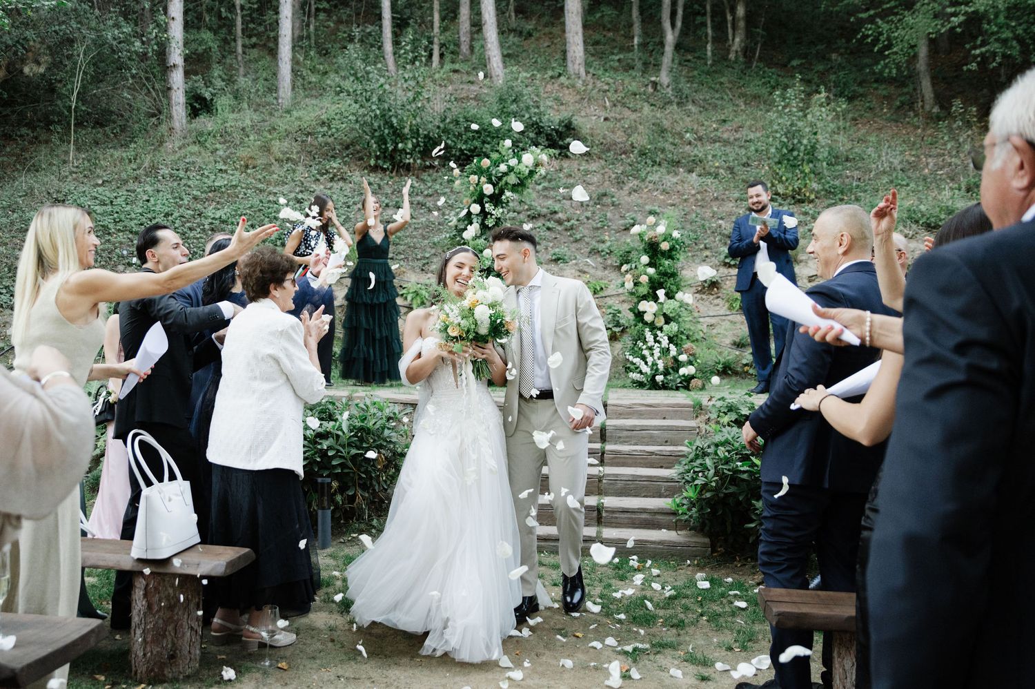 Wedding party and guests mingle in a scenic woodland setting during an outdoor celebration.