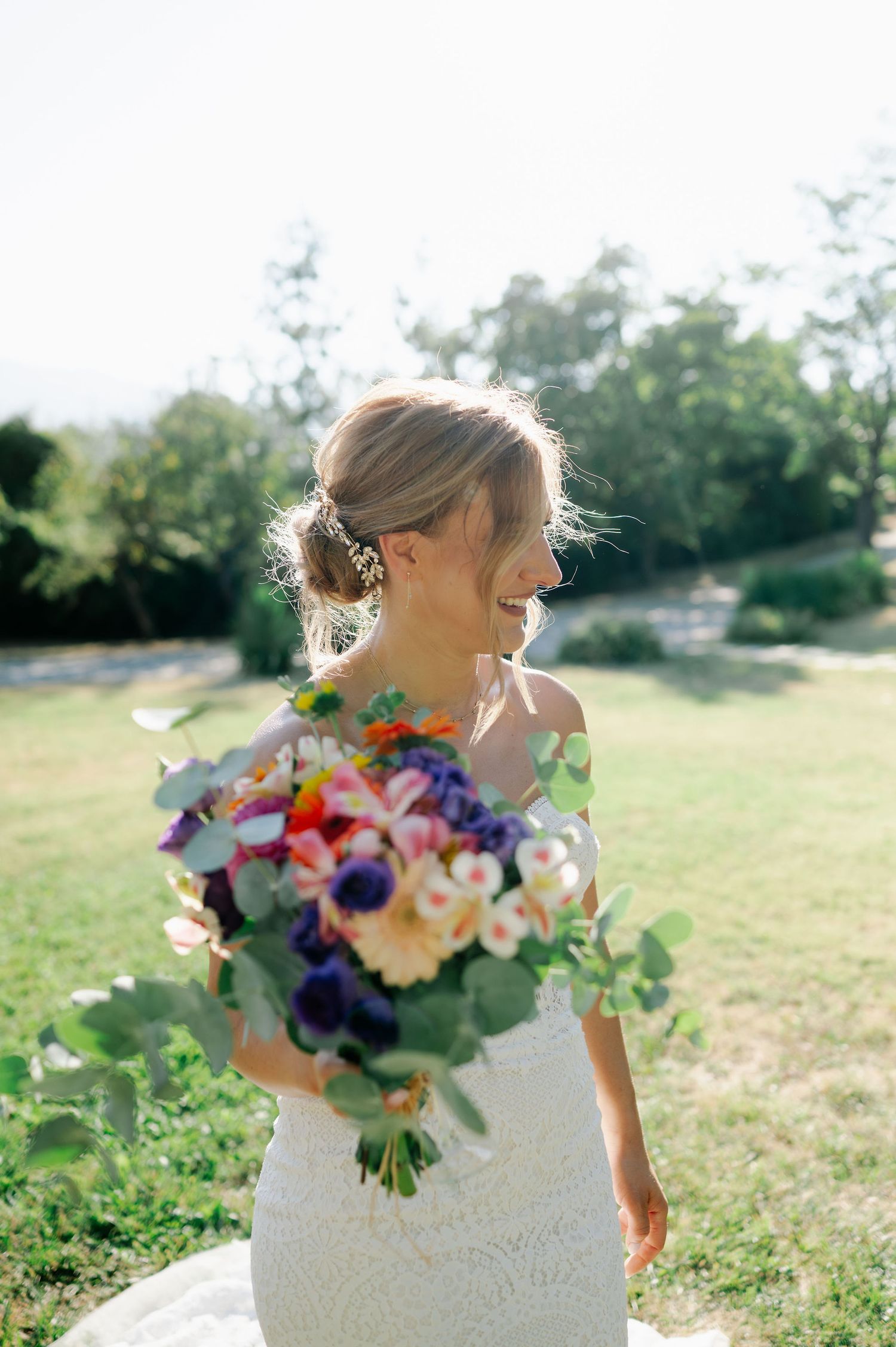 Person in white wedding dress holds a vibrant bouquet with purple and orange flowers in a sunny field.