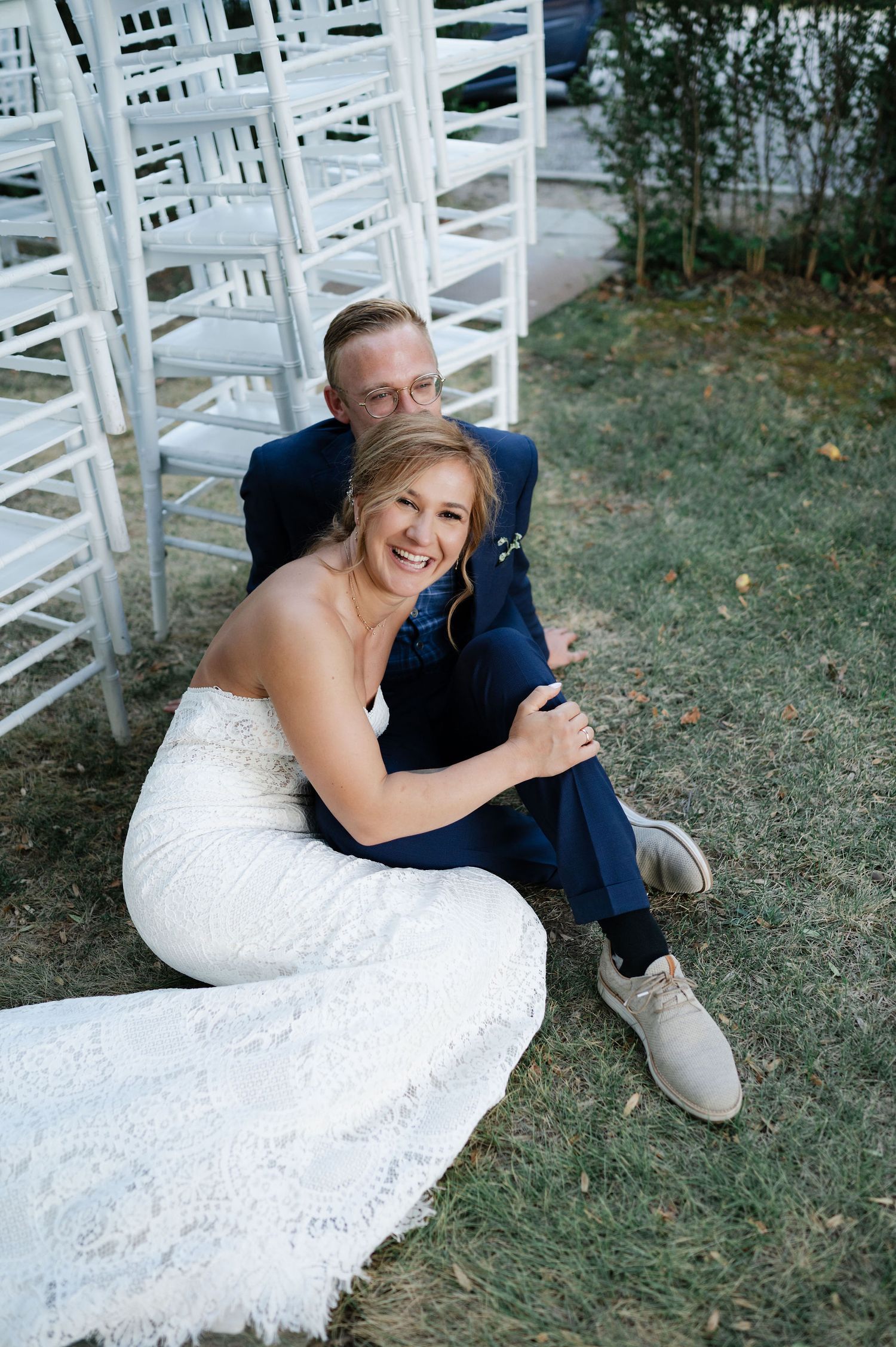 A bride in lace gown smiles while sitting on grass near white ceremony chairs.