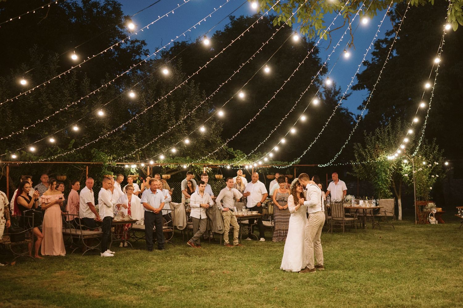Evening wedding reception under canopy of string lights with guests gathered around.