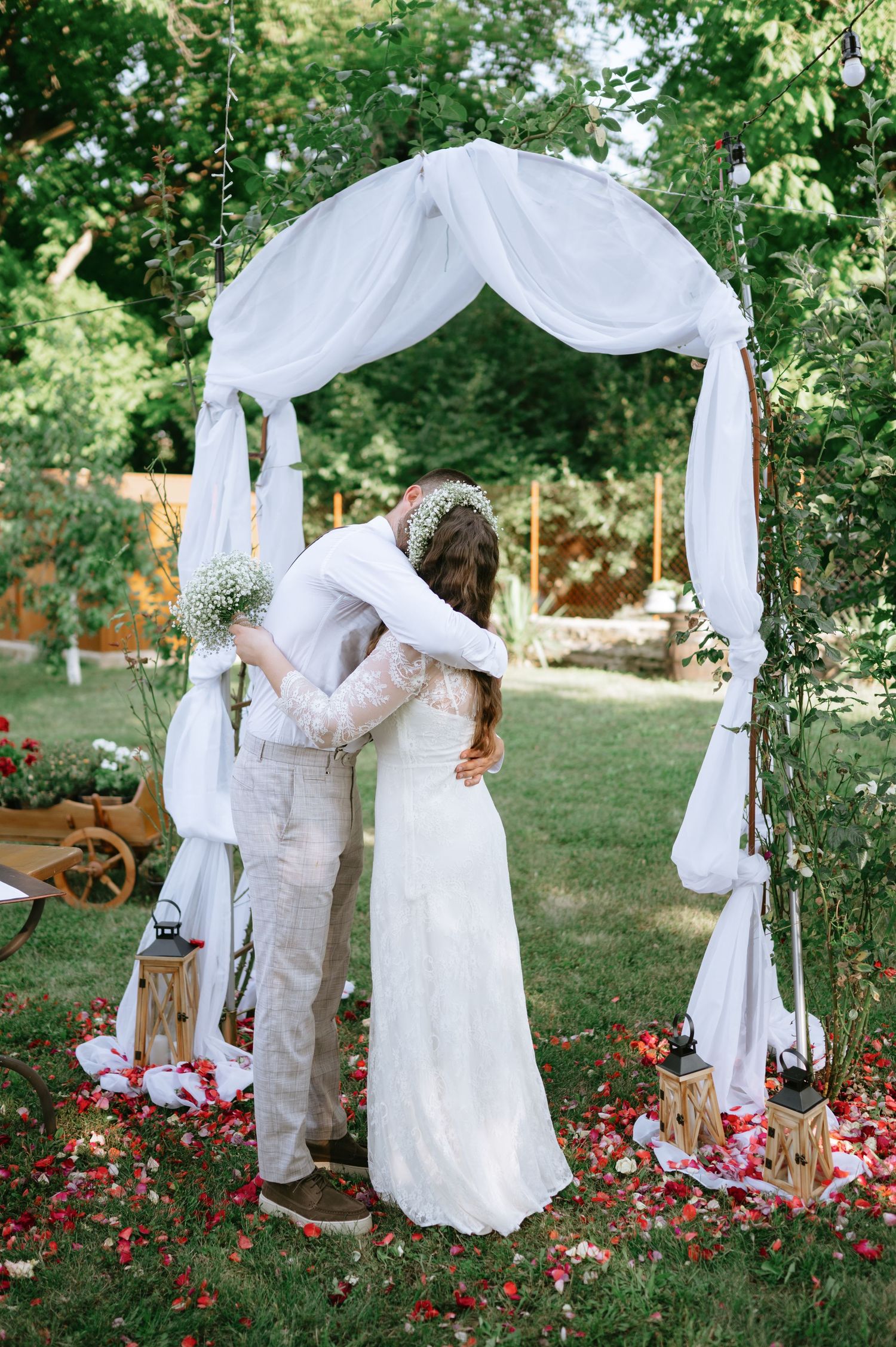 A romantic wedding moment captured under a white fabric arch with red flowers below.