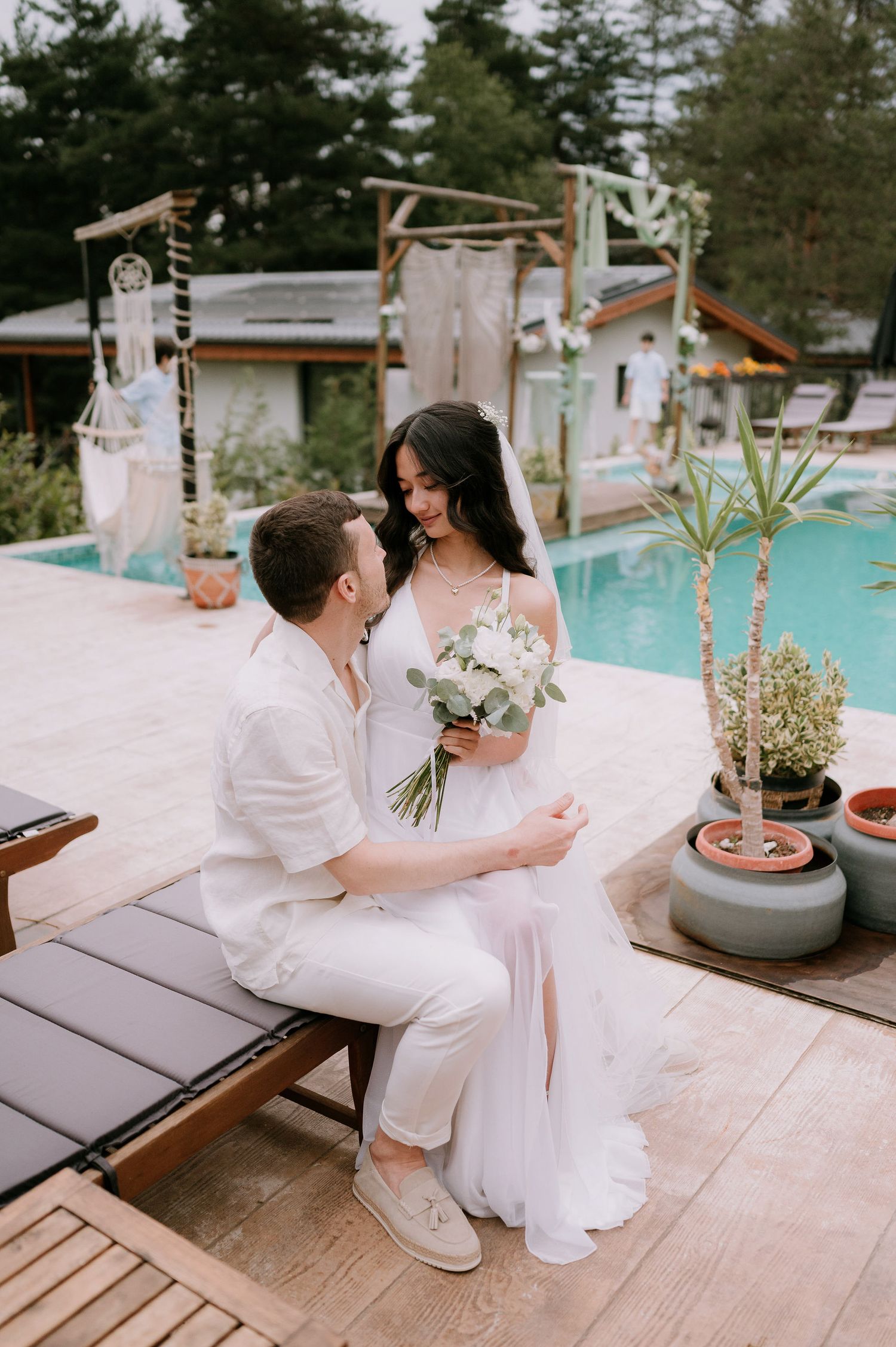 Wedding couple shares tender moments on wooden deck with pool and greenery in background.