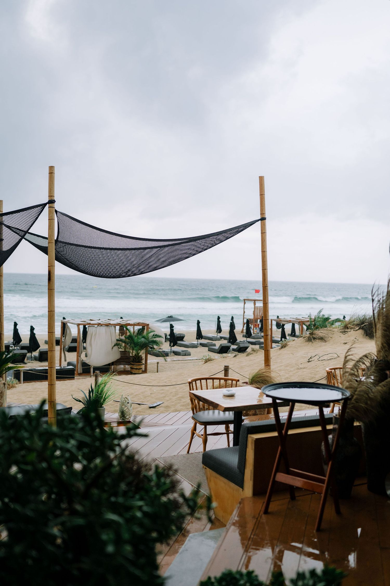 Coastal wedding venue with wooden furniture and black mesh canopies on a cloudy beach day.