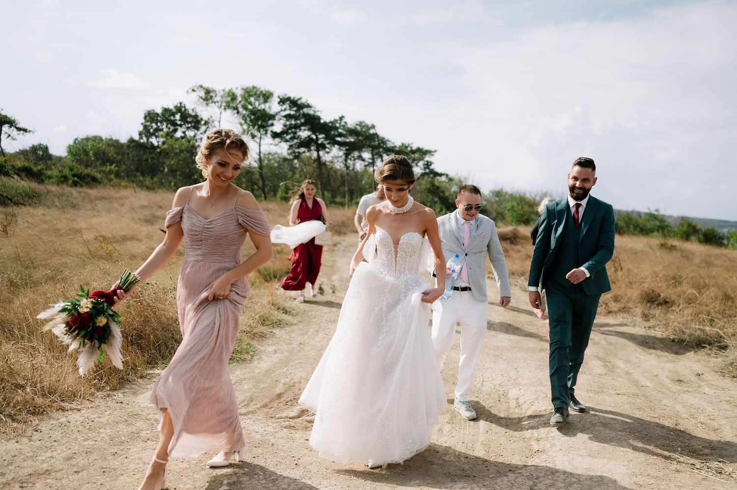 Wedding party members walking along a scenic dirt path on a sunny day.