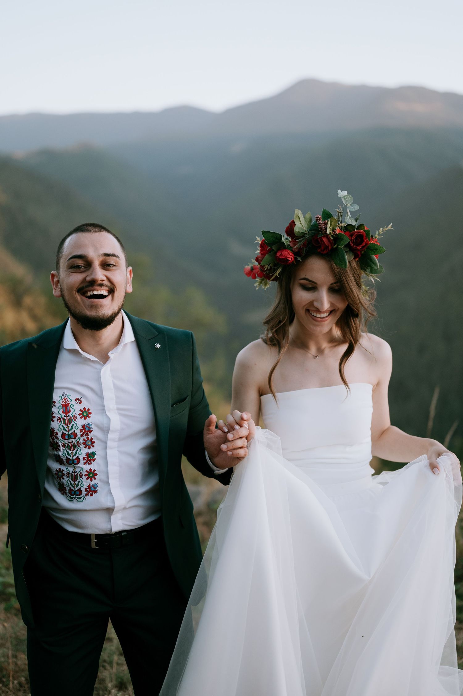 A couple in wedding attire shares joyful moments against a mountainous backdrop during sunset.