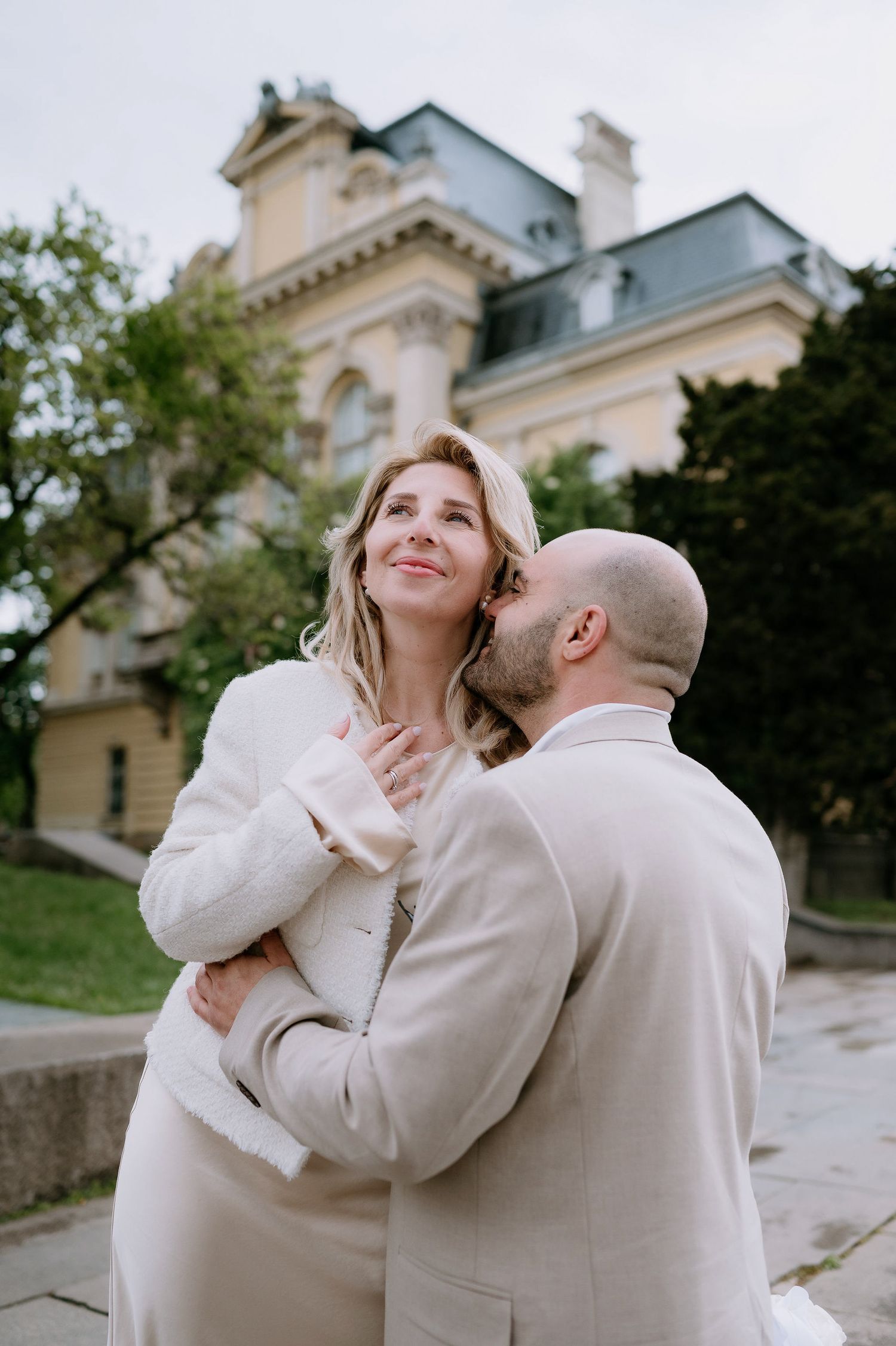 A romantic couple in white clothing shares a tender moment near an ornate building.