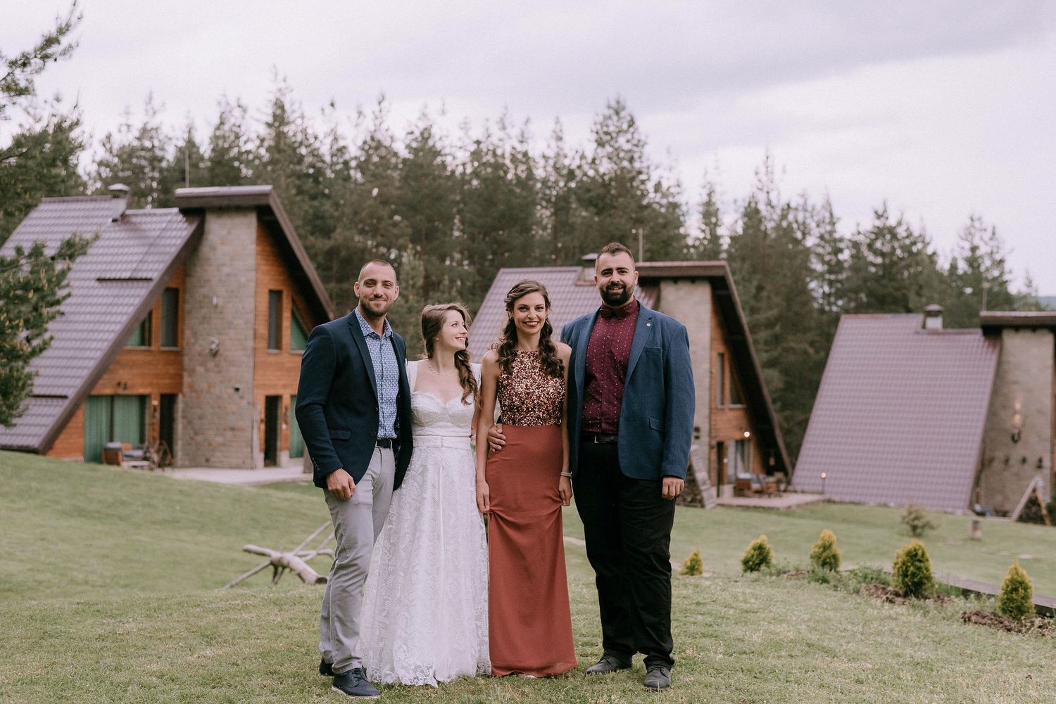 Wedding party poses in front of rustic A-frame cabins surrounded by pine trees.