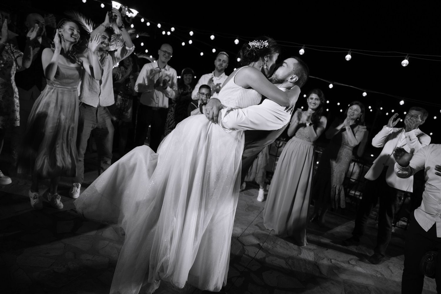 A couple shares a romantic dance at their outdoor evening wedding reception under string lights while guests look on.