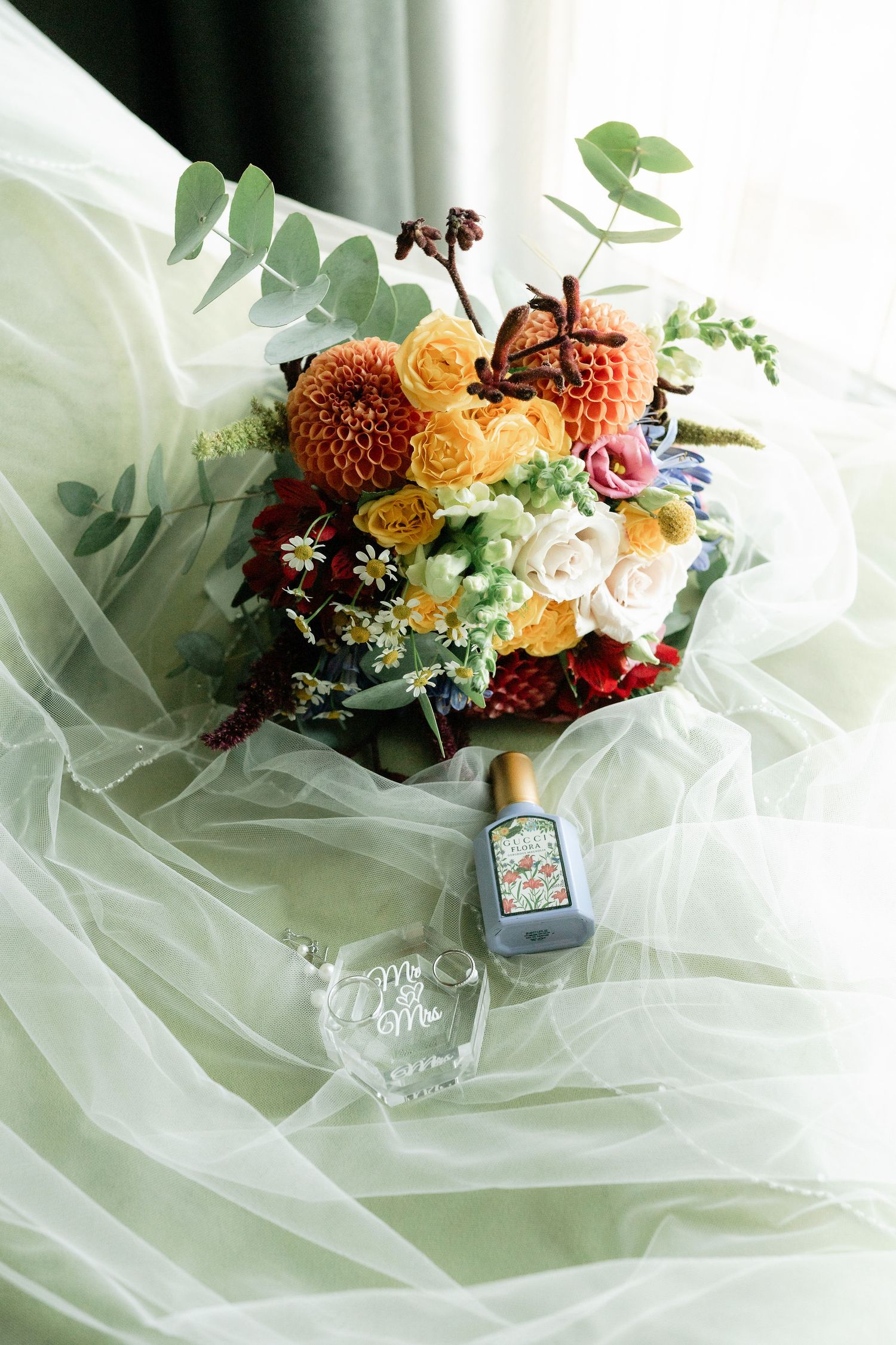 A delicate bouquet of orange, pink and burgundy flowers with greenery rests on white tulle fabric.