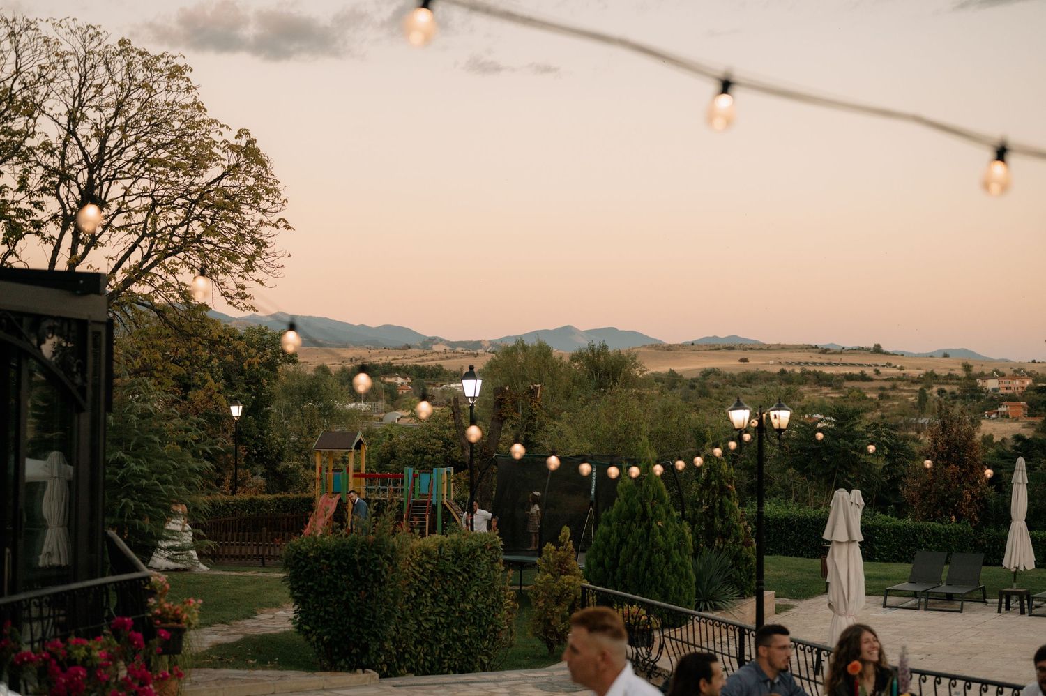 String lights illuminate an outdoor patio at dusk with rolling hills and mountains visible in the background.
