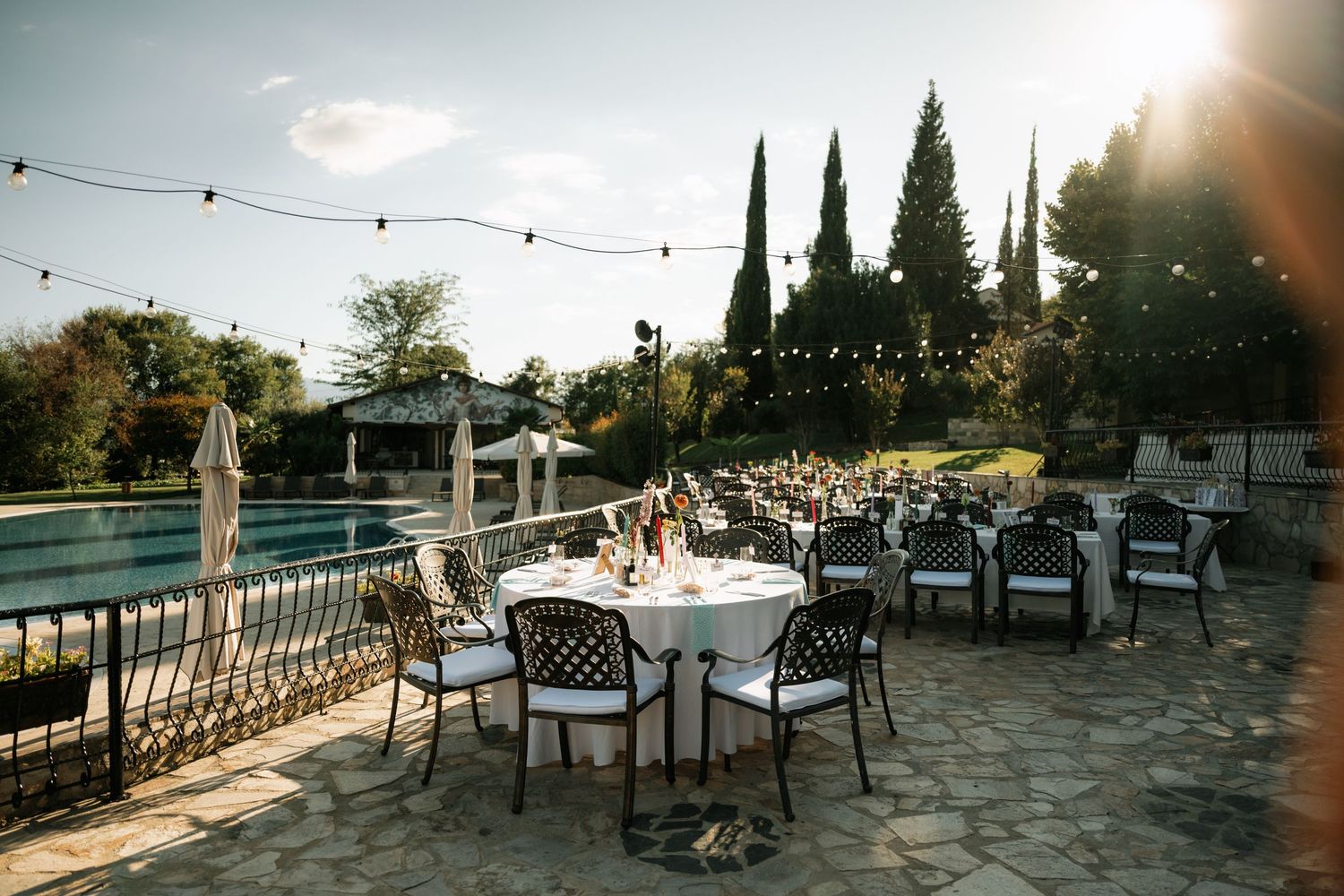 String lights illuminate an outdoor patio dining area with tables and chairs at sunset.