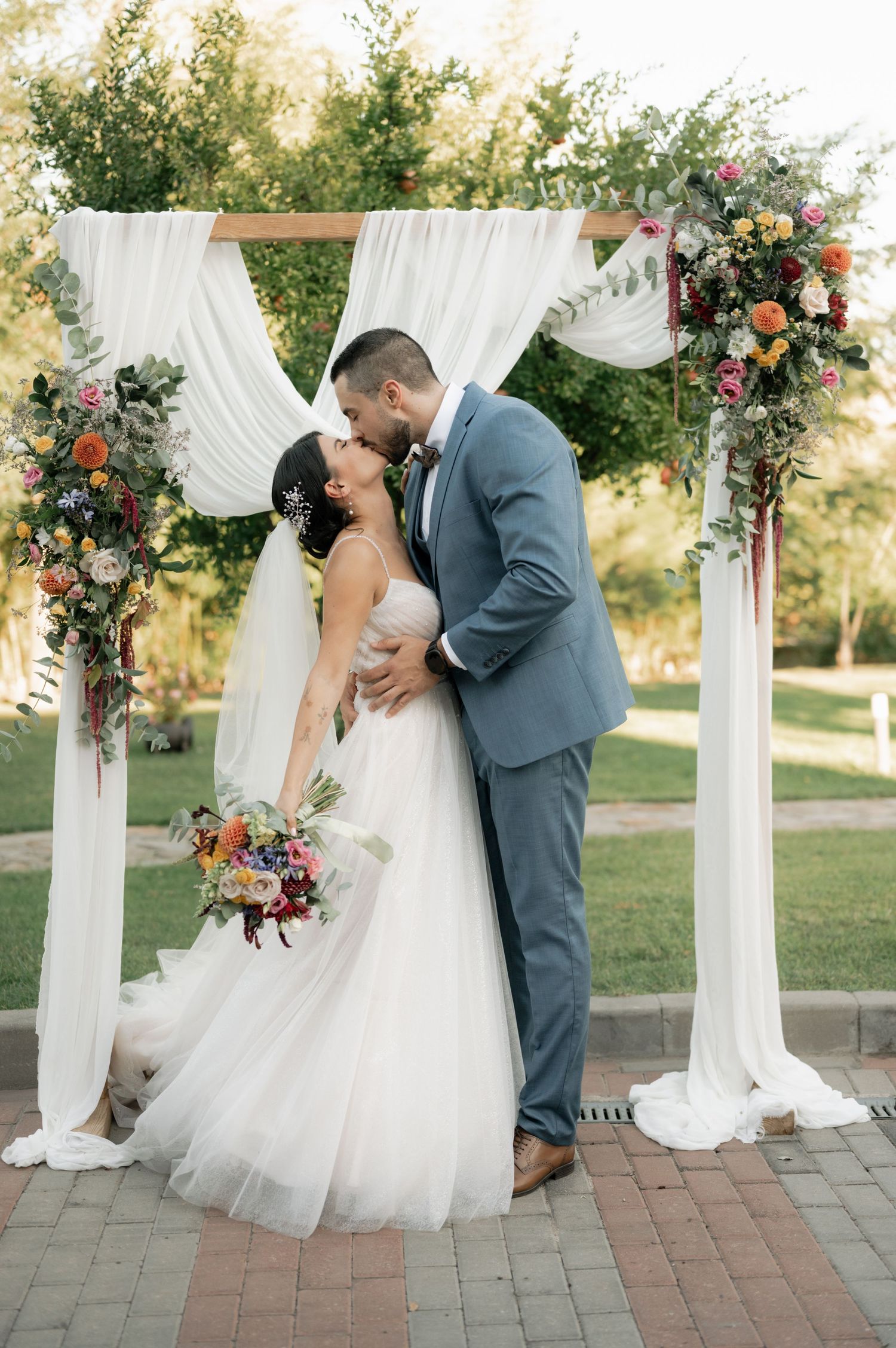 Romantic outdoor wedding ceremony with white draped arch, floral arrangements and brick pathway at sunset.
