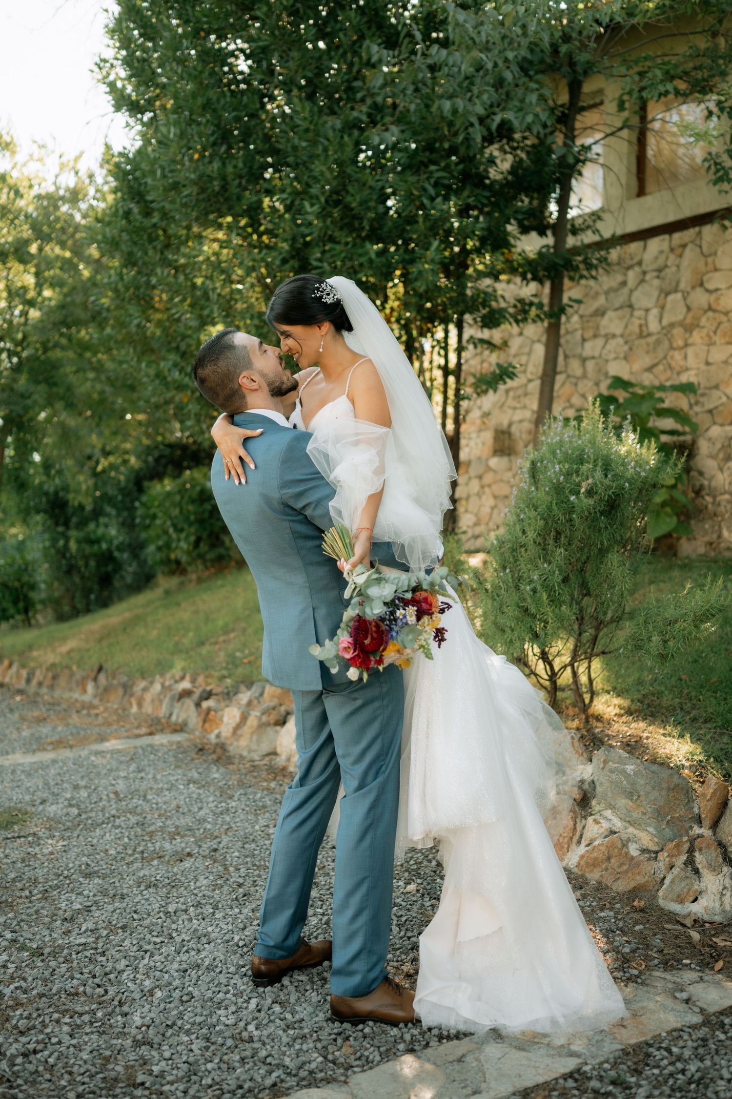 A couple shares a romantic moment on a gravel path by a stone wall, with the bride holding a burgundy bouquet.