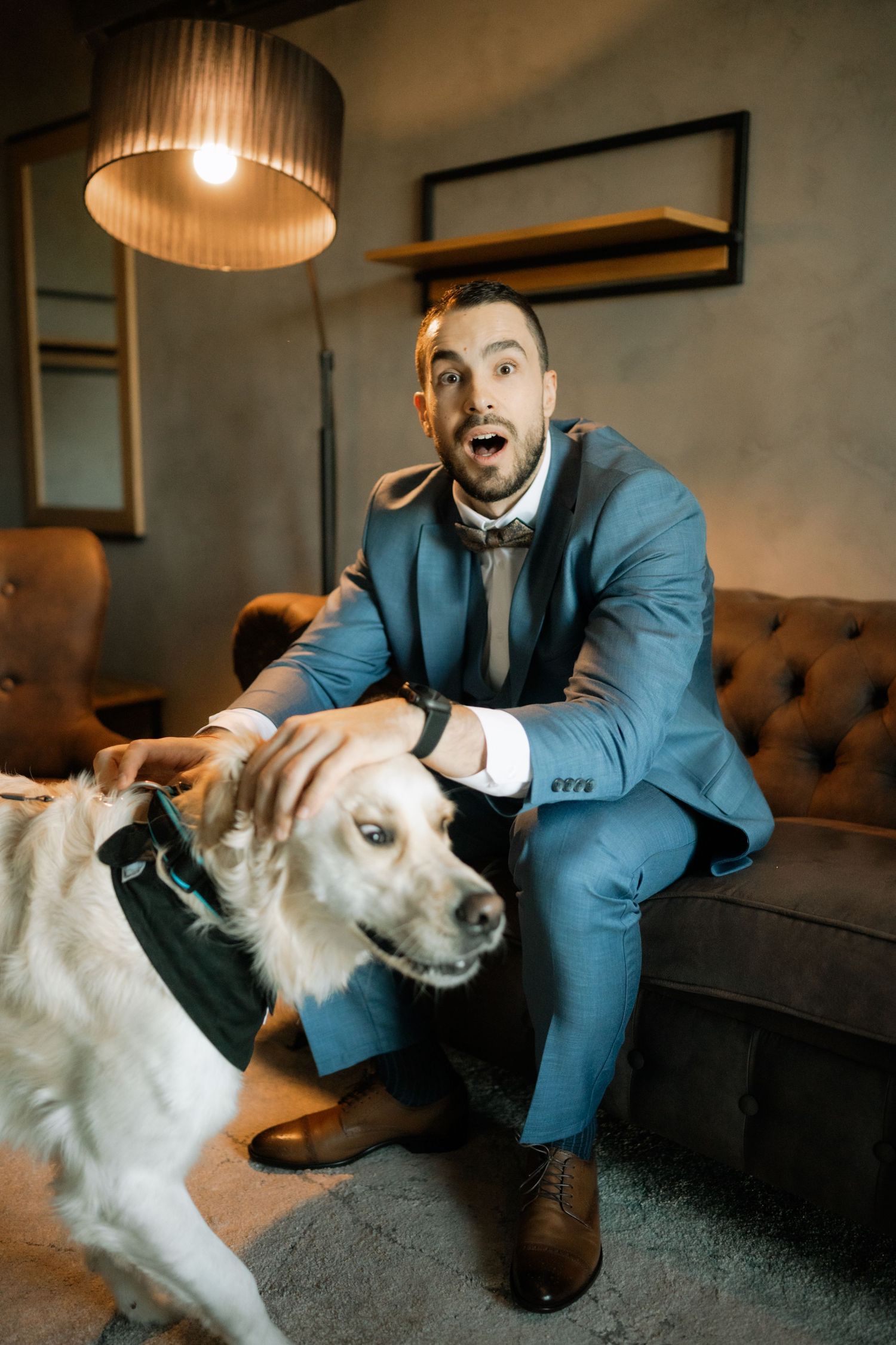 A person in a blue suit crouches down to pet a white dog on a brown couch in a dimly lit room.