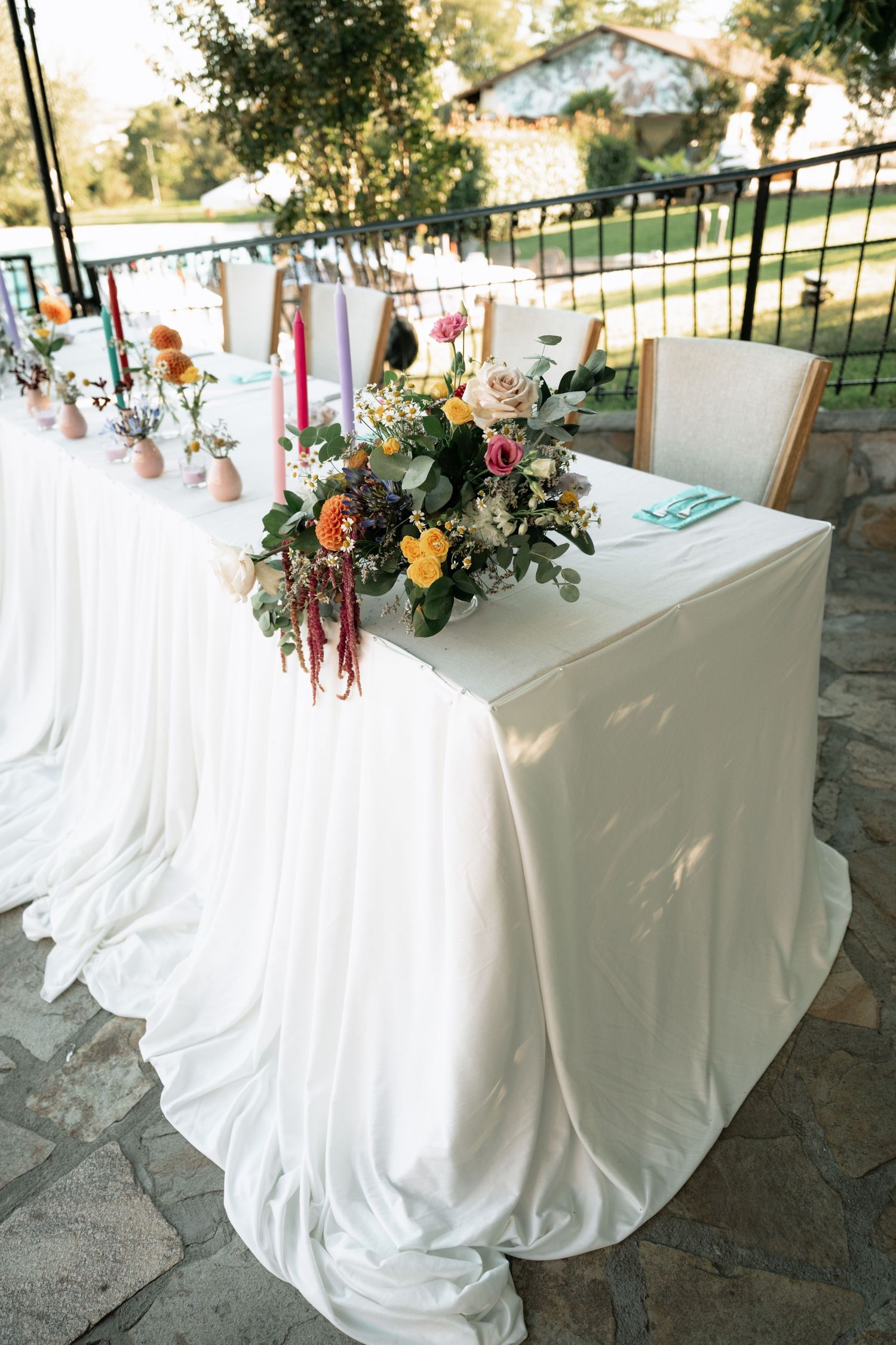 Elegant white tablecloths drape over reception tables decorated with floral arrangements and greenery in an outdoor venue setting.