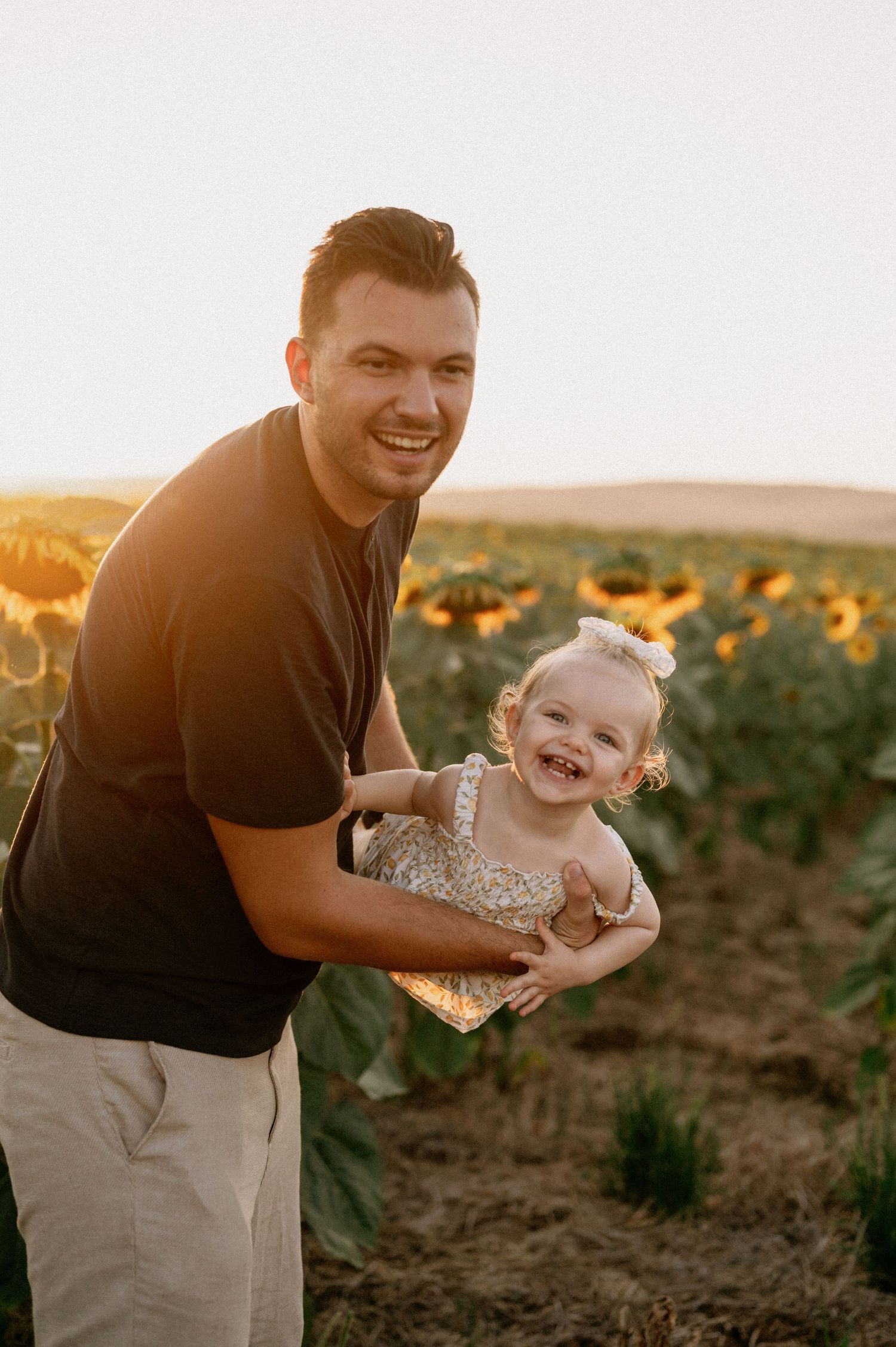 Smiling man holding a laughing baby girl in a sunflower field during golden hour sunset.