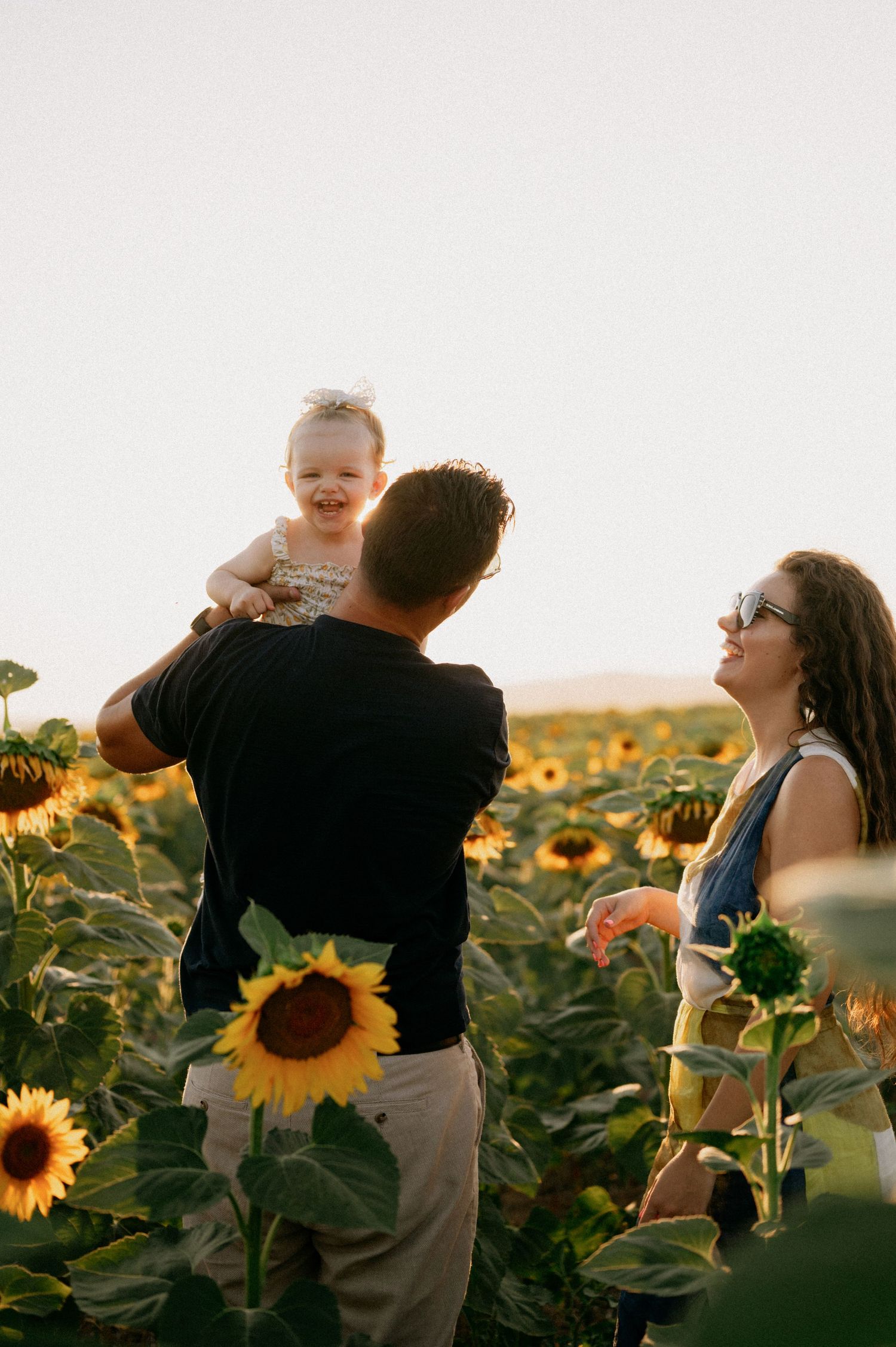 Family enjoying golden hour in a sunflower field, father lifting baby girl while mother smiles nearby.