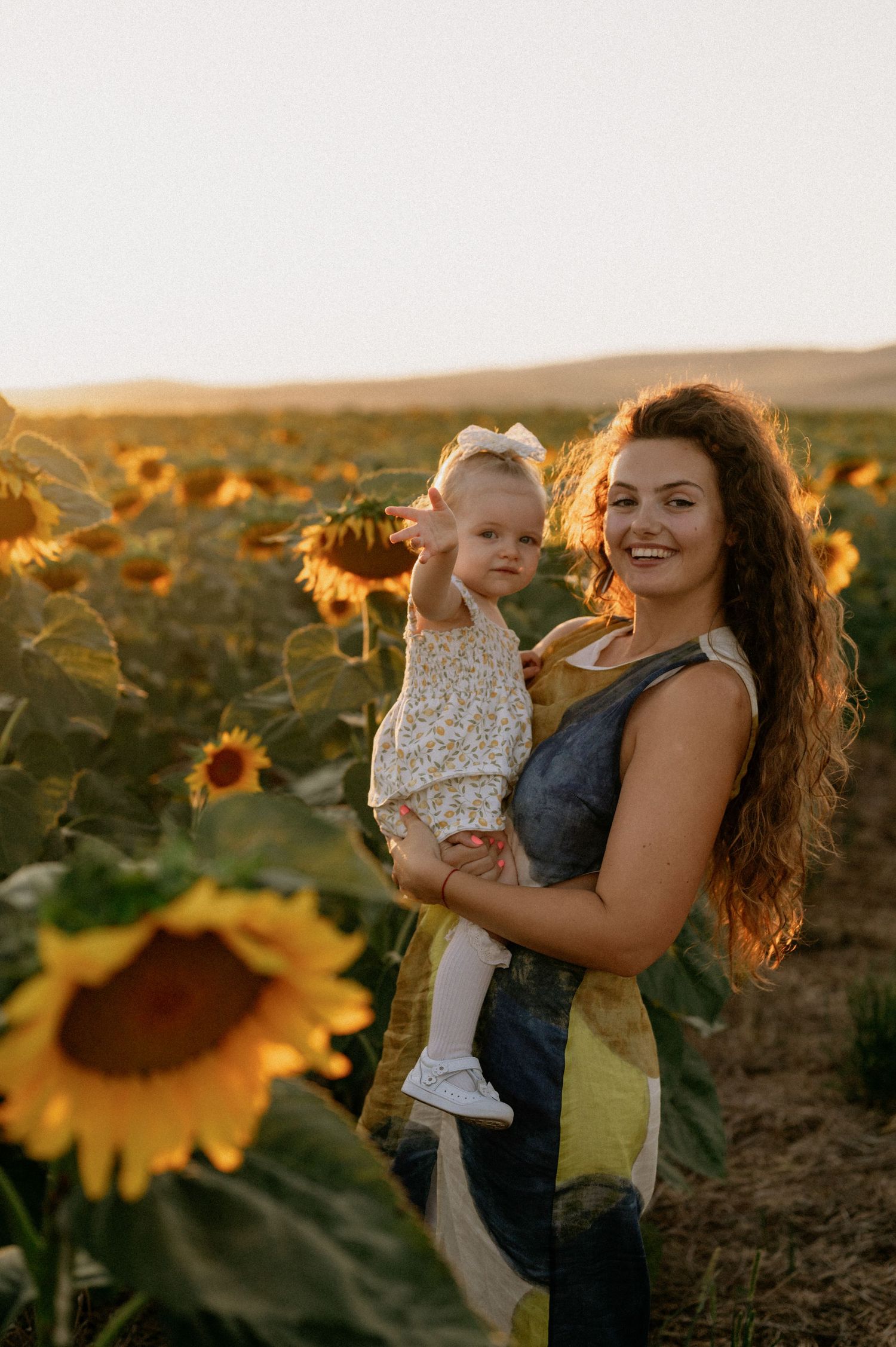 Smiling woman holding a baby girl in a sunflower field at golden hour sunset.