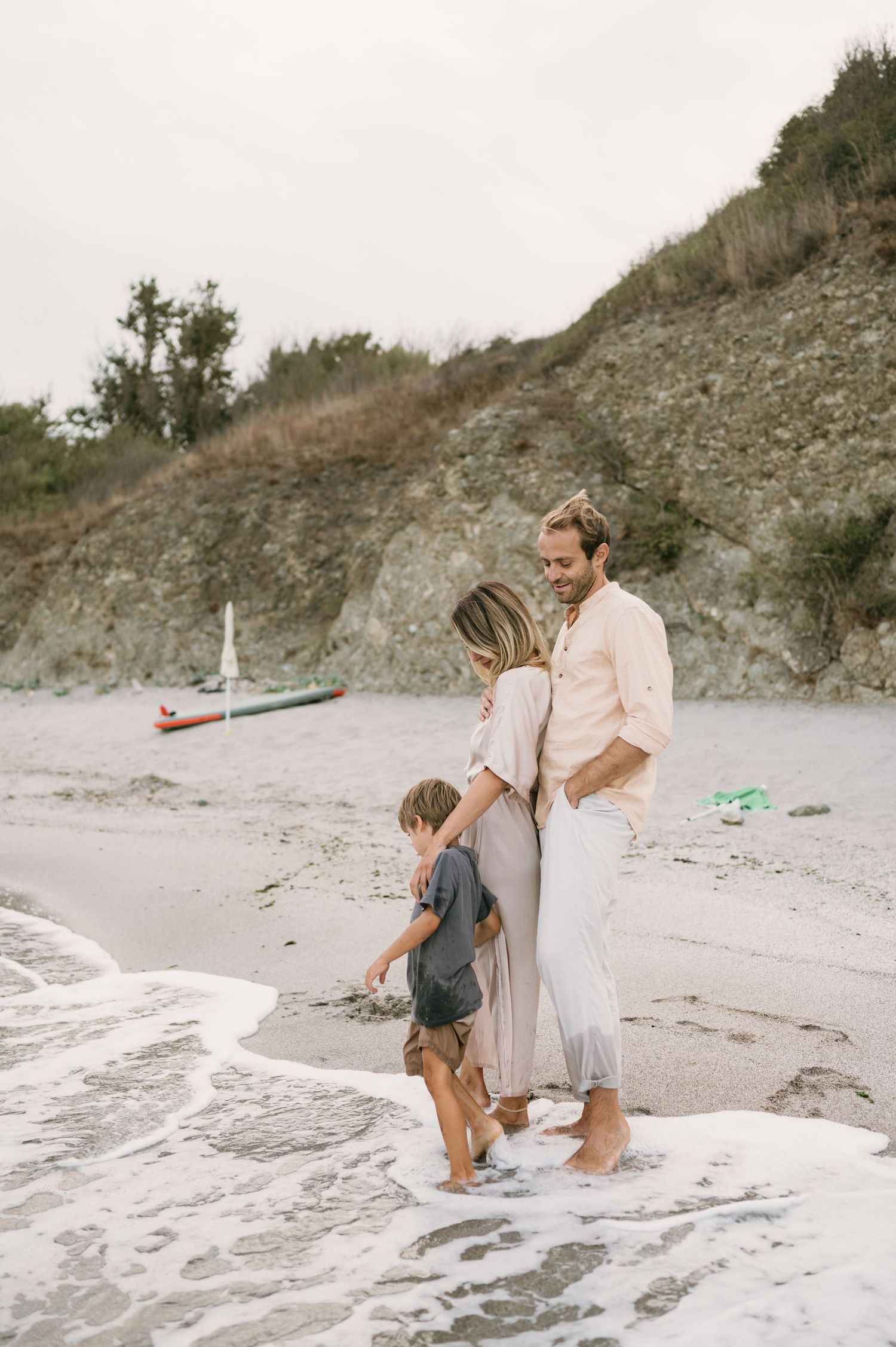 Family of three walking along sandy beach shore with rocky cliffs and boats in background on overcast day.