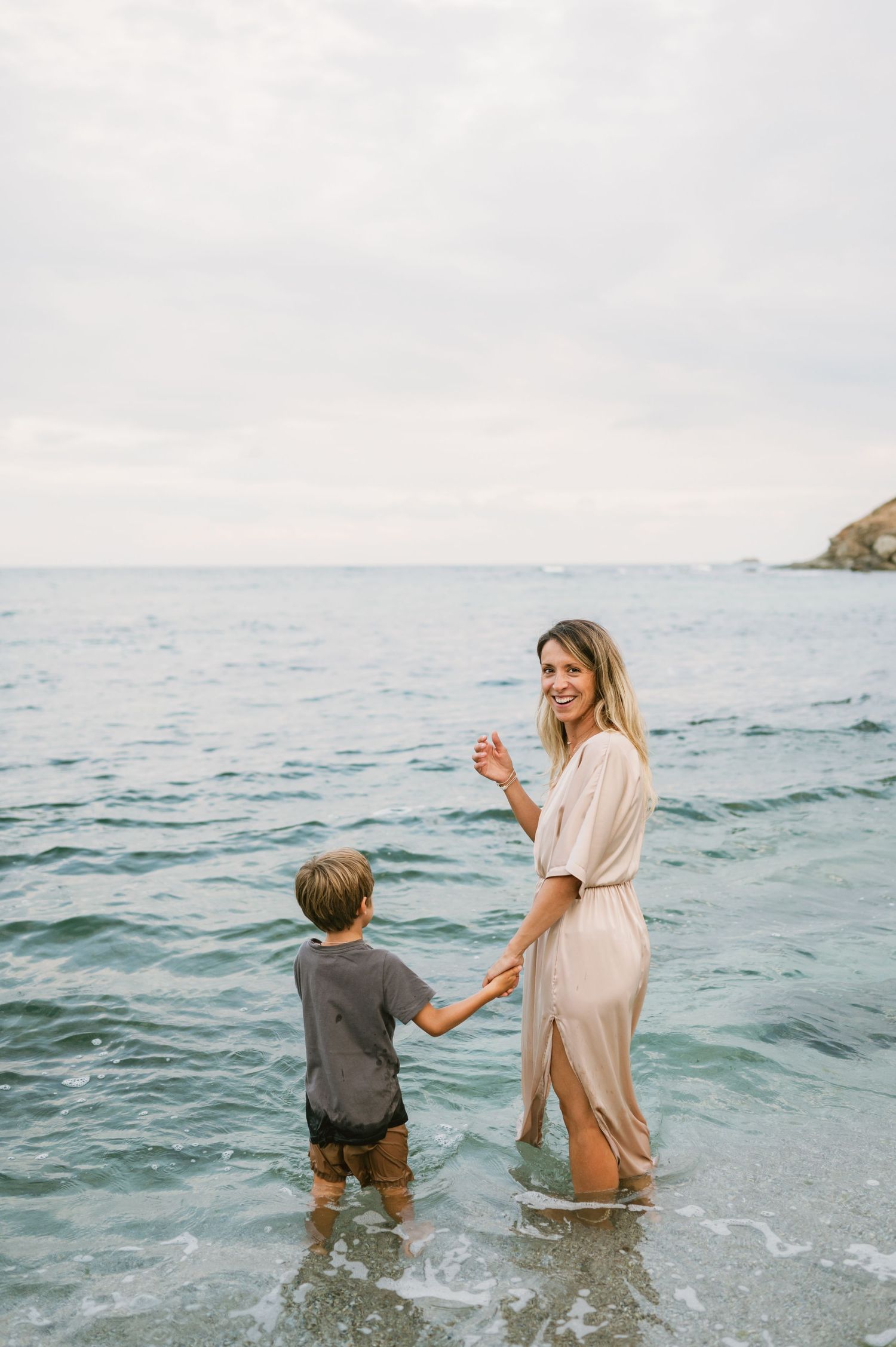 A smiling woman in a beige dress holds hands with a young boy wading in shallow ocean waves at the beach.