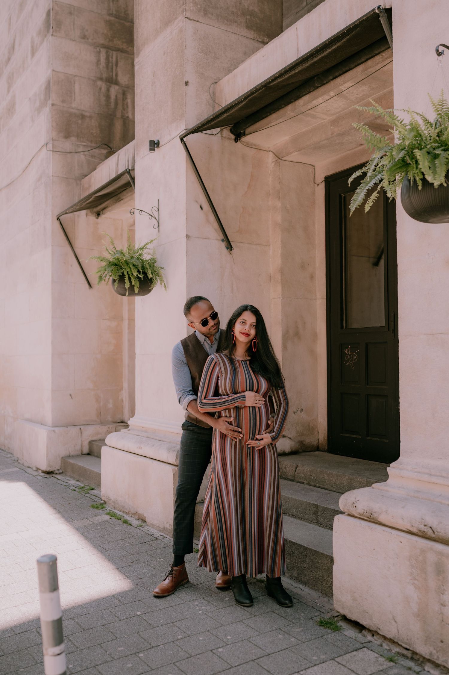 A couple poses outside a stone building, the woman in a striped maxi dress cradling her baby bump.