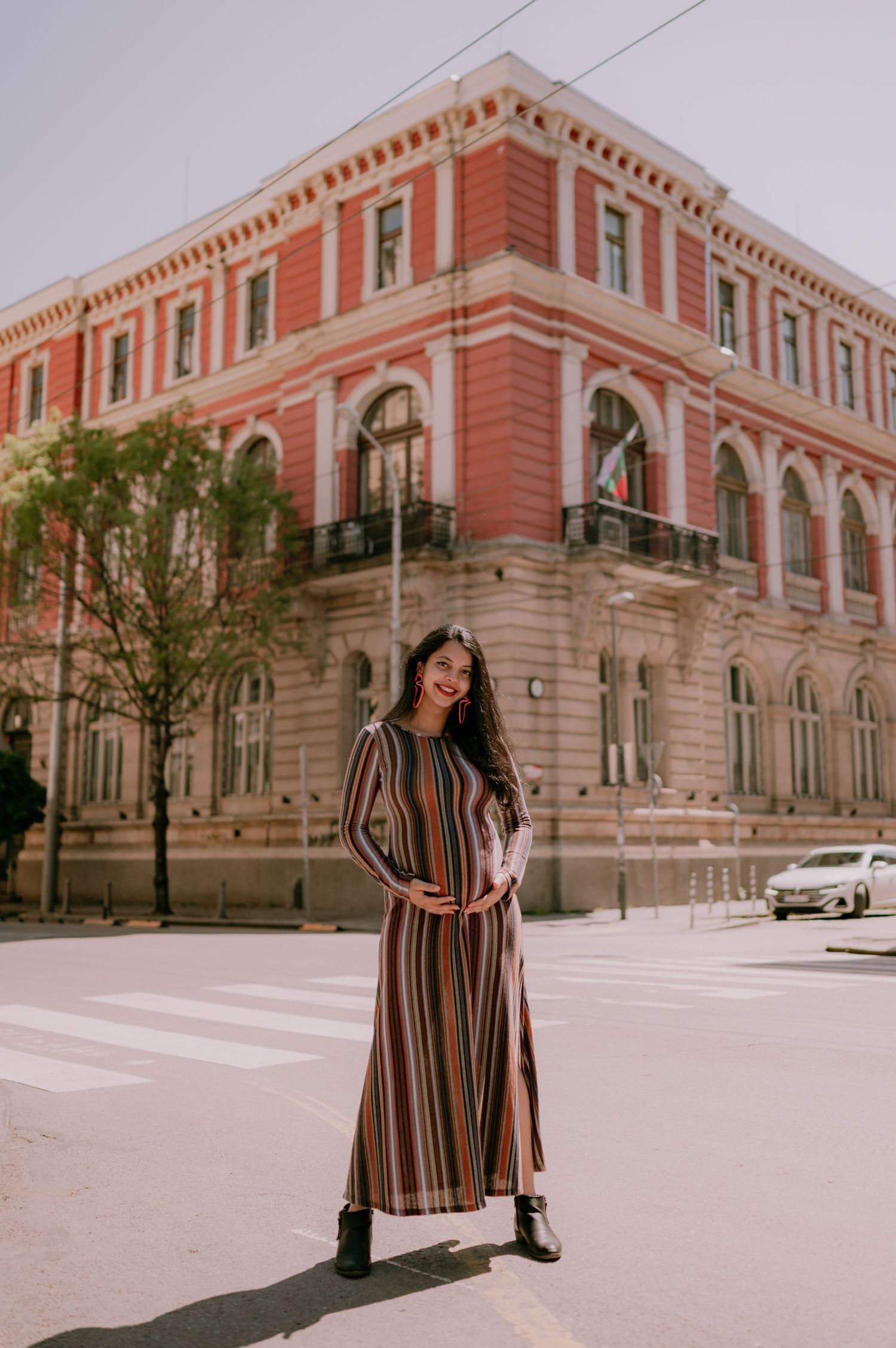 Woman in striped maxi dress and black boots posing on street corner near historic red building.