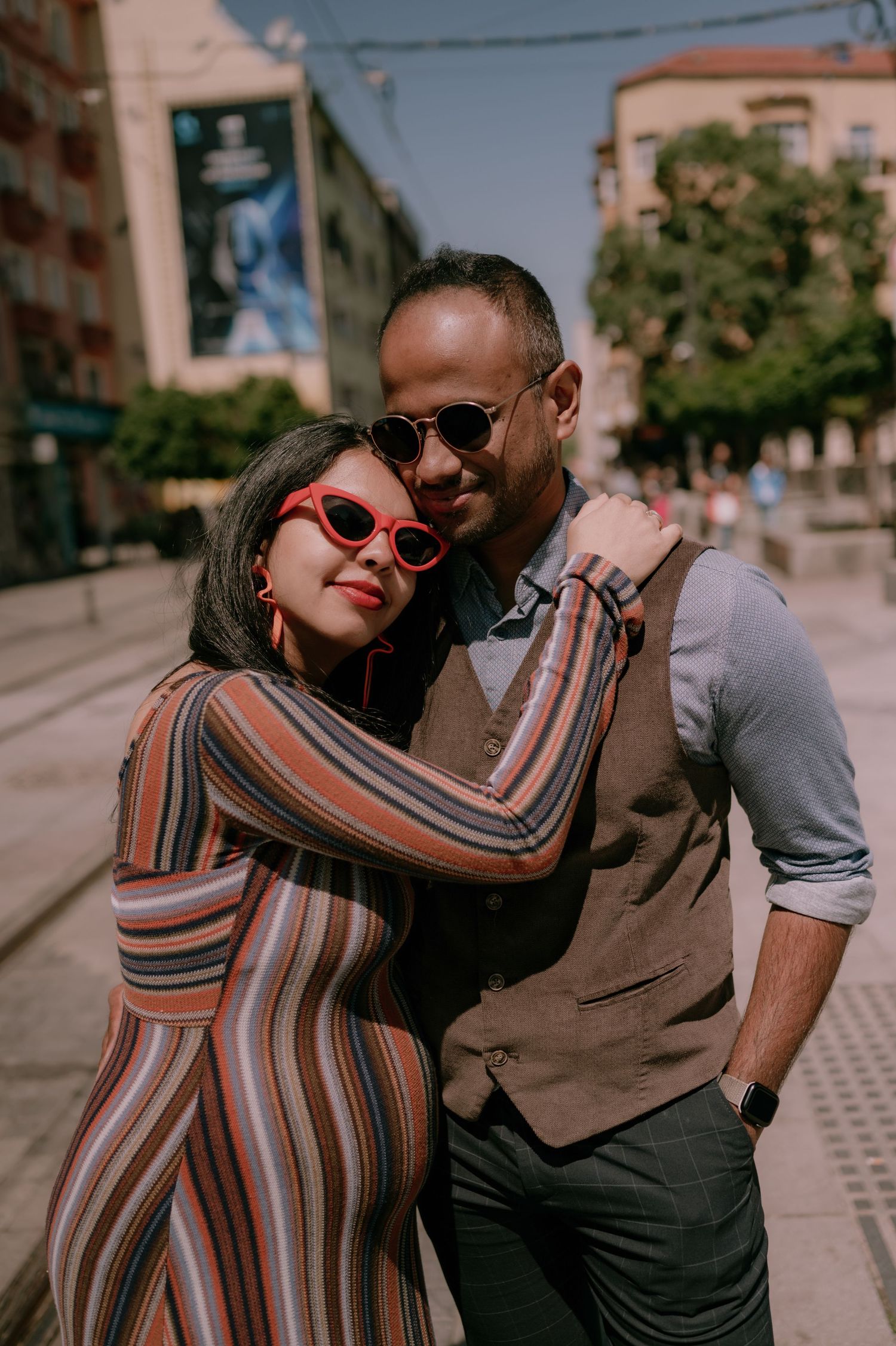 A stylish couple embraces on a sunny urban street, woman in striped jumpsuit with red sunglasses, man in vest and dress shirt.