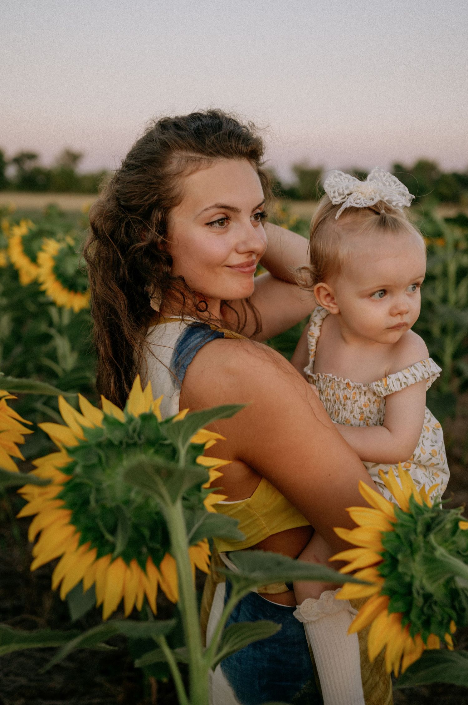 Young woman holding baby girl in sunflower field at sunset, both wearing summer outfits in golden light.