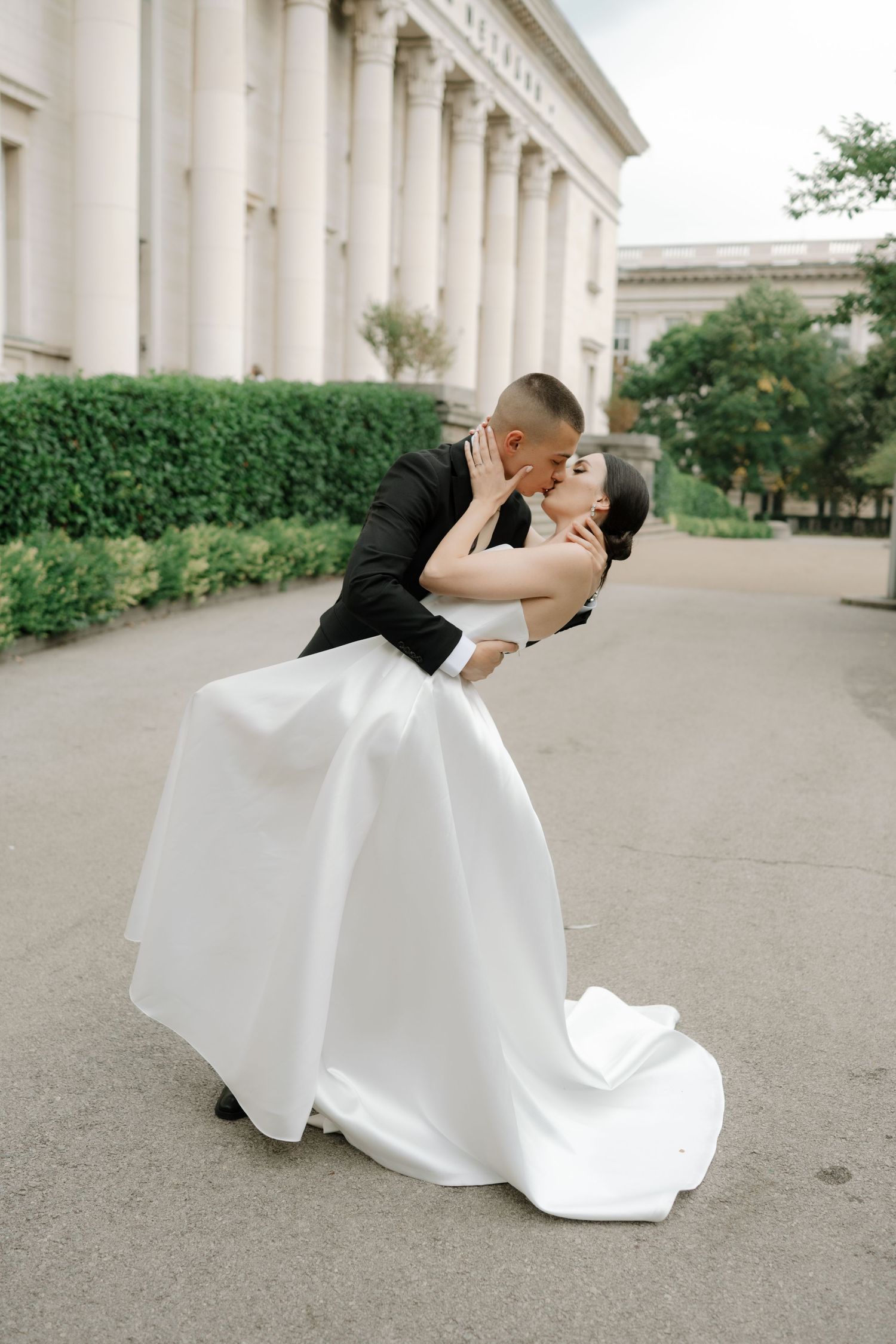 A romantic wedding couple shares a kiss in front of a grand columned building with a flowing white dress and classic tuxedo.