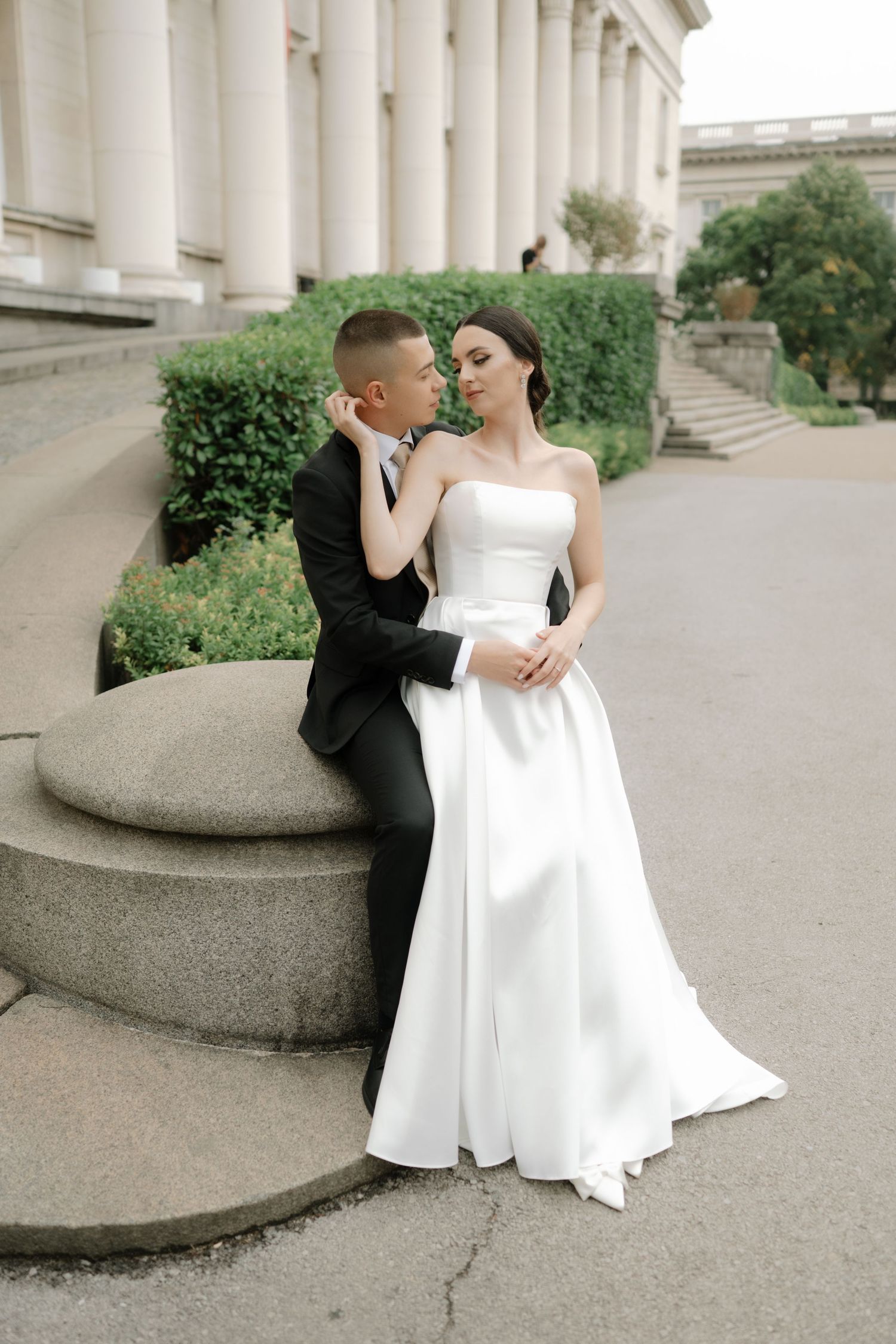 A couple in formal wedding attire pose elegantly against classic stone columns and greenery.