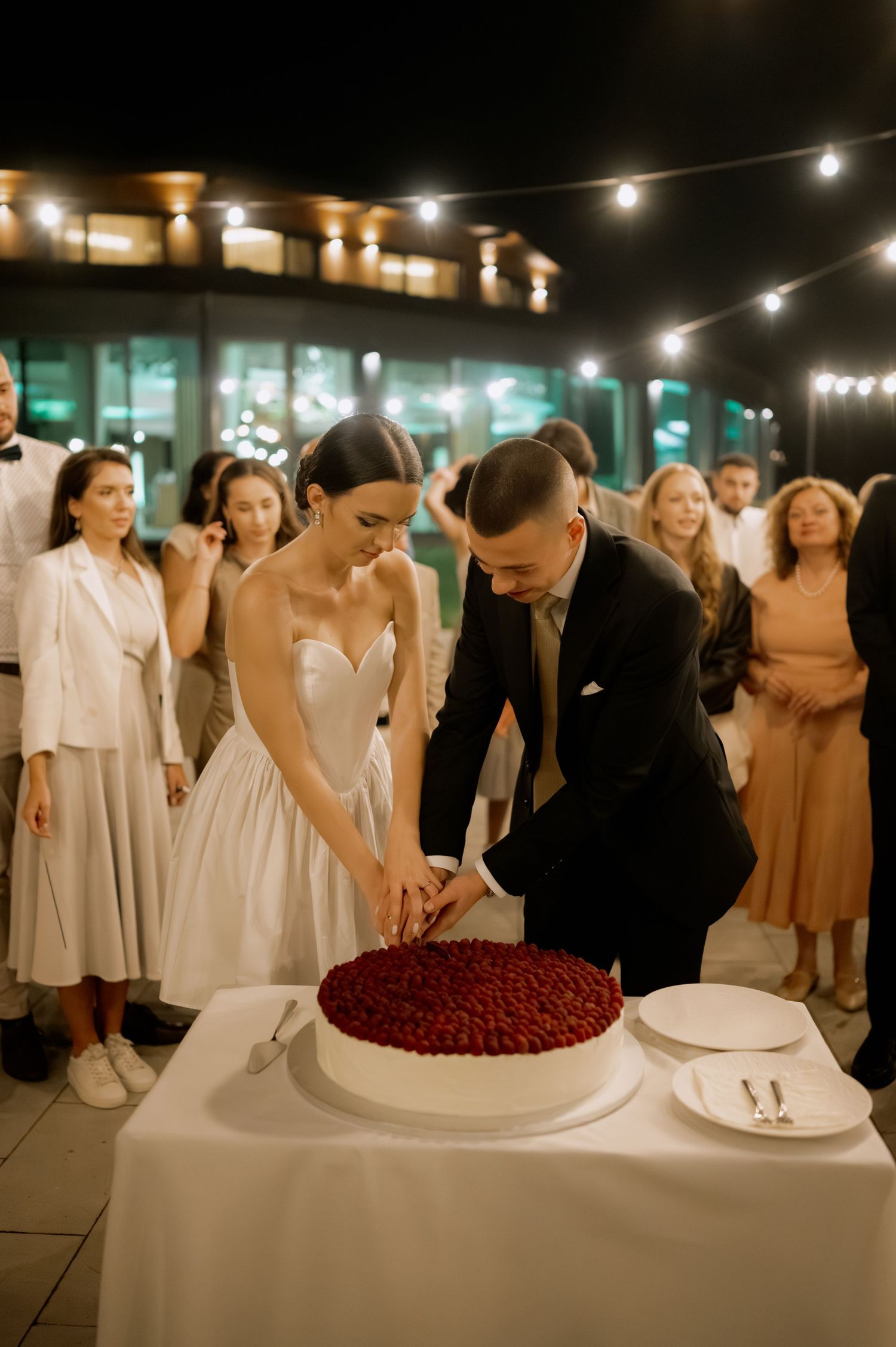 Wedding guests gathered around a dessert table with a red berry cake under string lights at night.