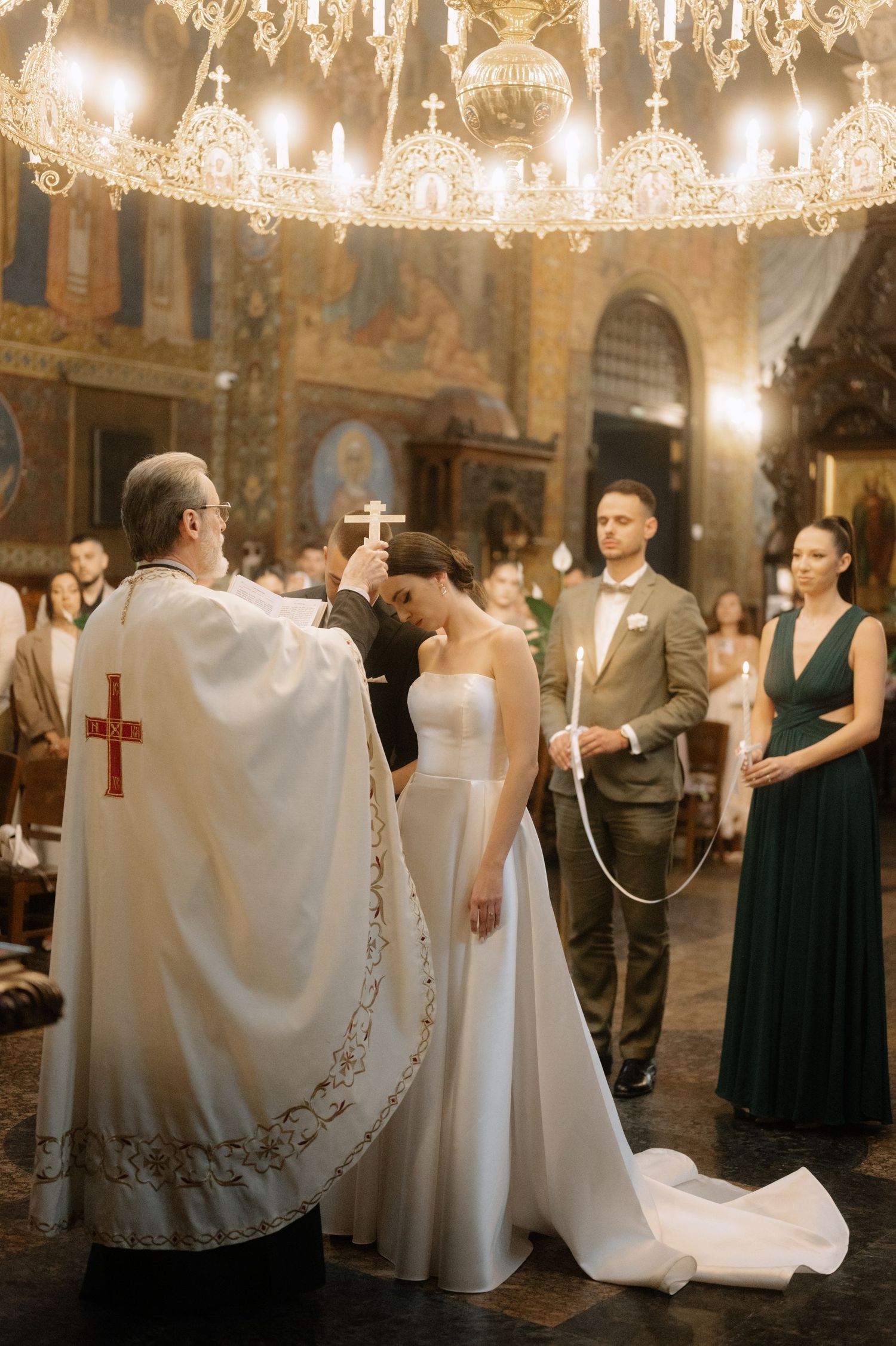 Wedding ceremony in an ornate church with crystal chandeliers showing guests in formal attire and white robes.