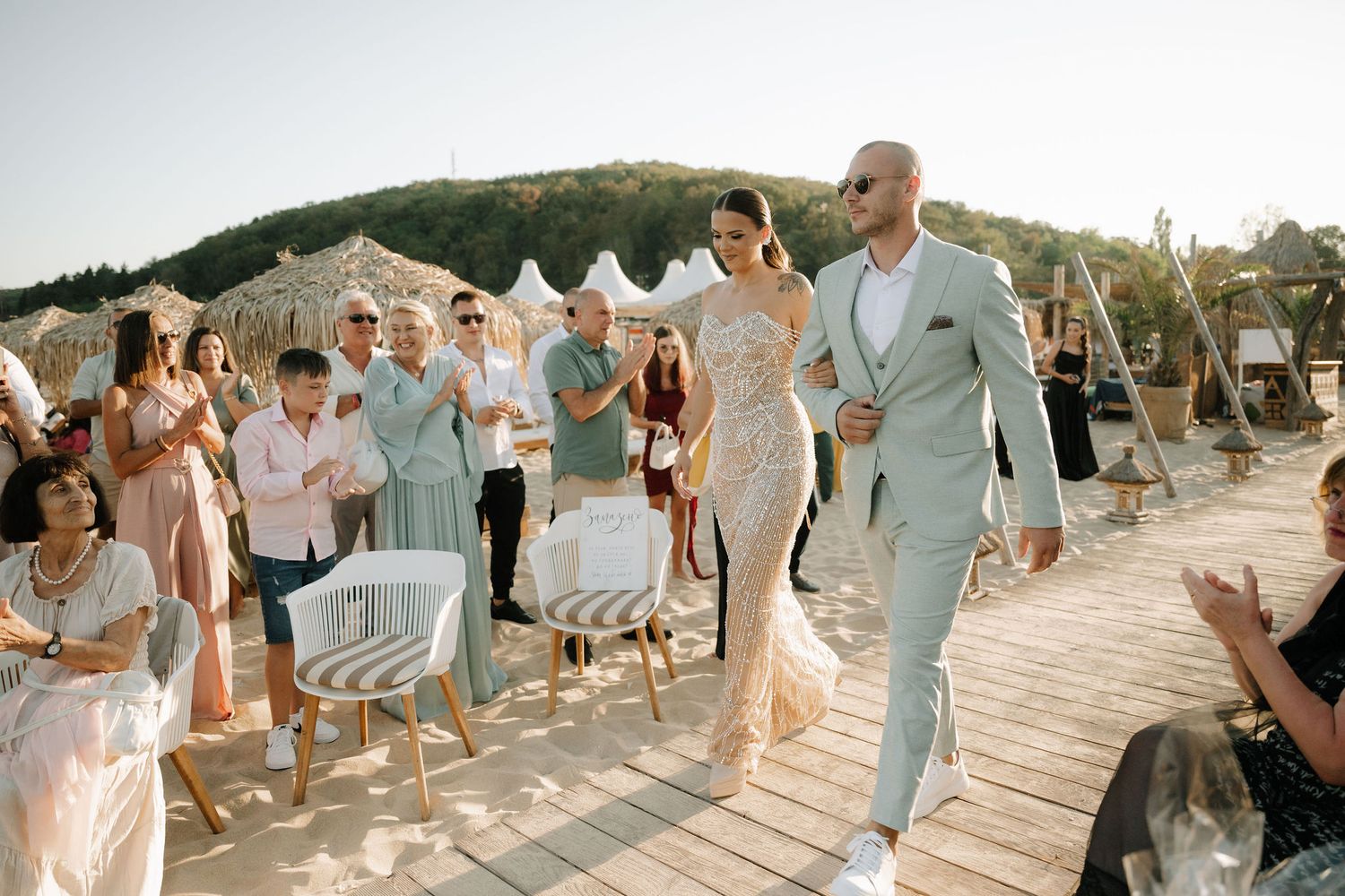 Wedding guests celebrate during a beachside ceremony on a wooden deck at sunset with white tents in the background.