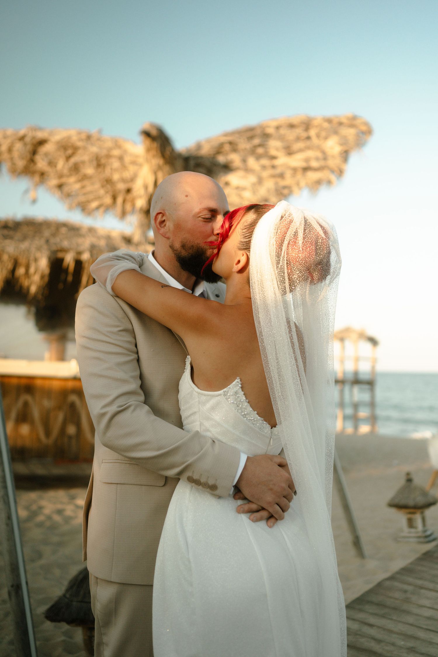 Romantic beach wedding sequence showing couple kissing at sunset with thatched umbrellas and ocean views in the background.