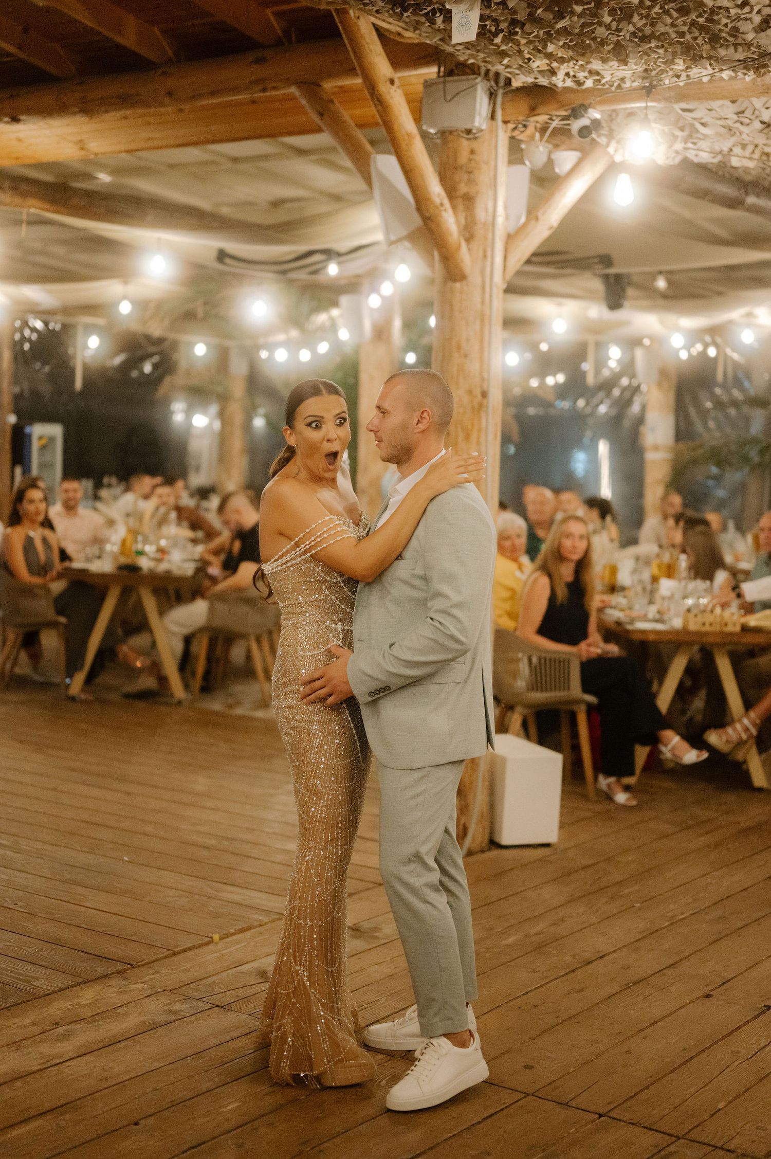 A couple shares a romantic dance together on a rustic wooden deck under twinkling string lights at an evening reception.