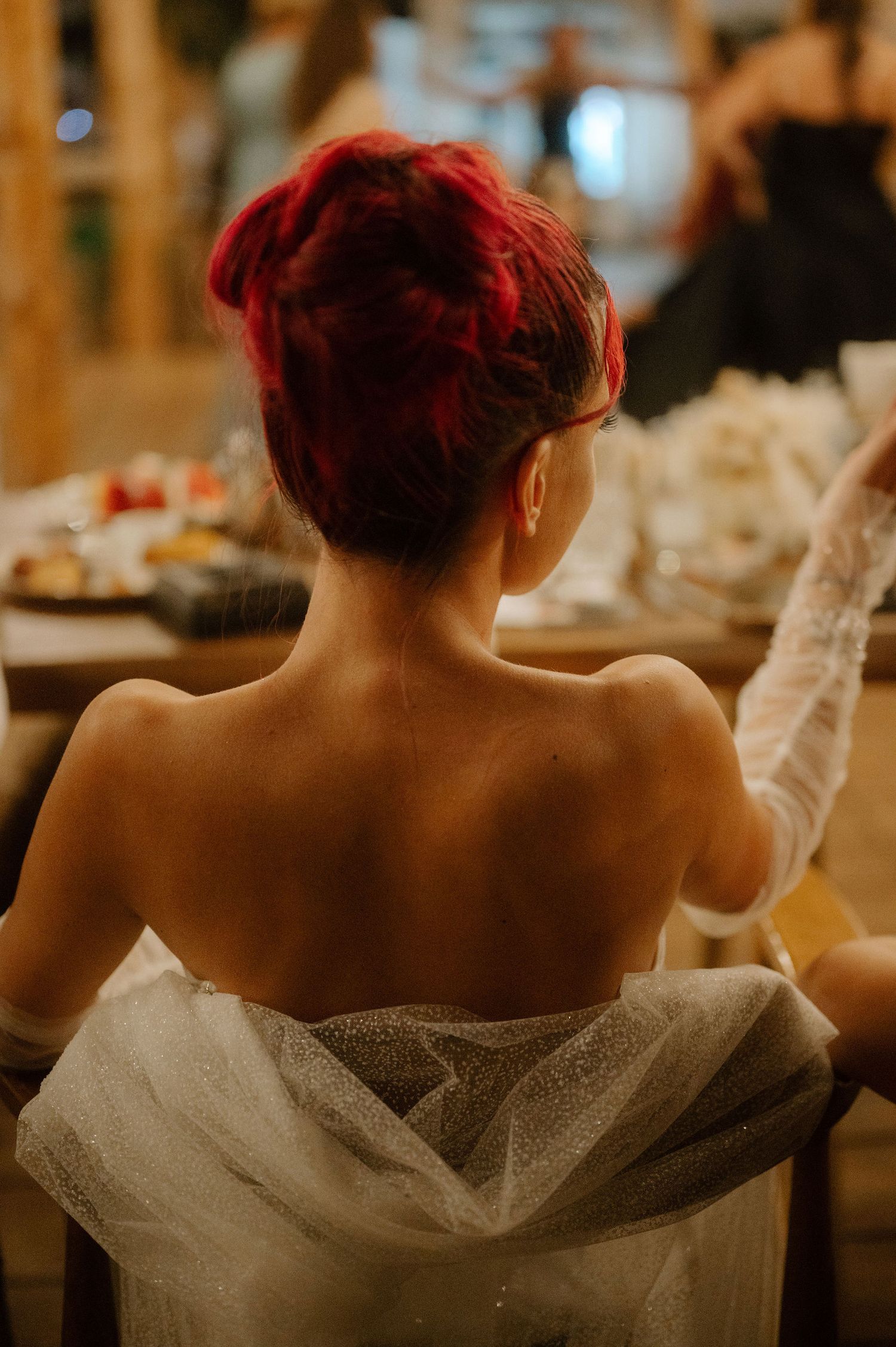 Back view of someone with vibrant red hair wearing a strapless white tulle dress in a warm indoor setting.