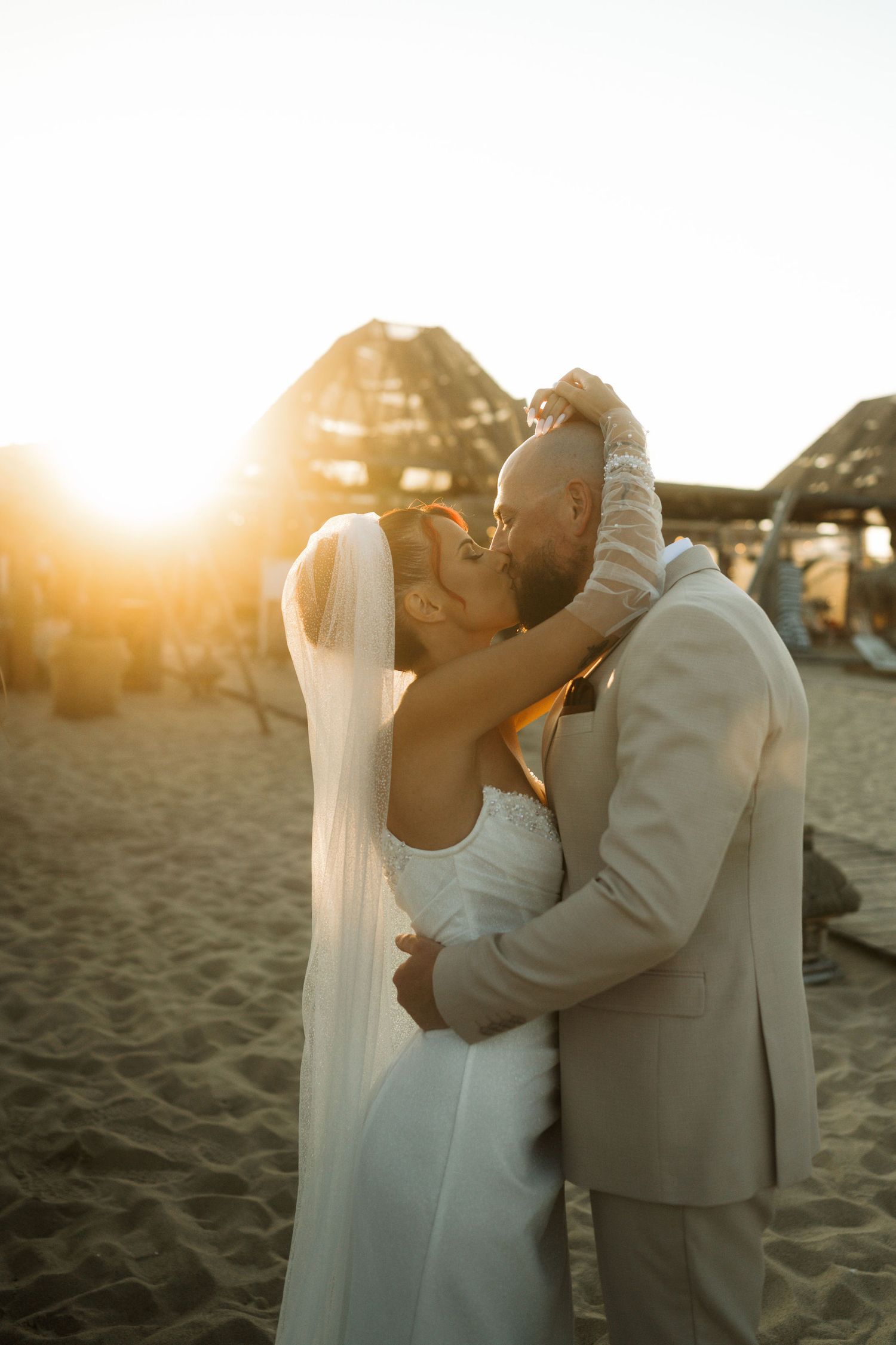 A romantic sunset beach wedding moment with couple embracing in formal attire as golden light glows behind them.