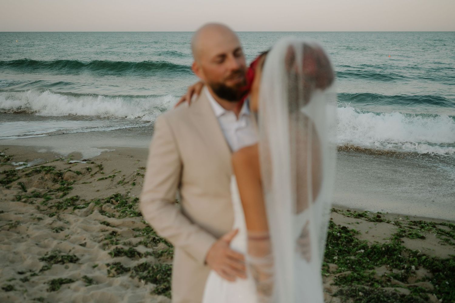 A couple in wedding attire share a romantic moment on a beach at sunset with ocean waves in the background.