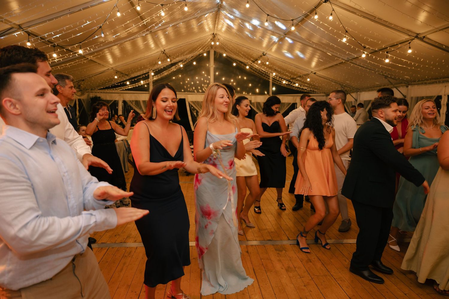 Wedding guests dance and celebrate together under a white tent at an evening reception.