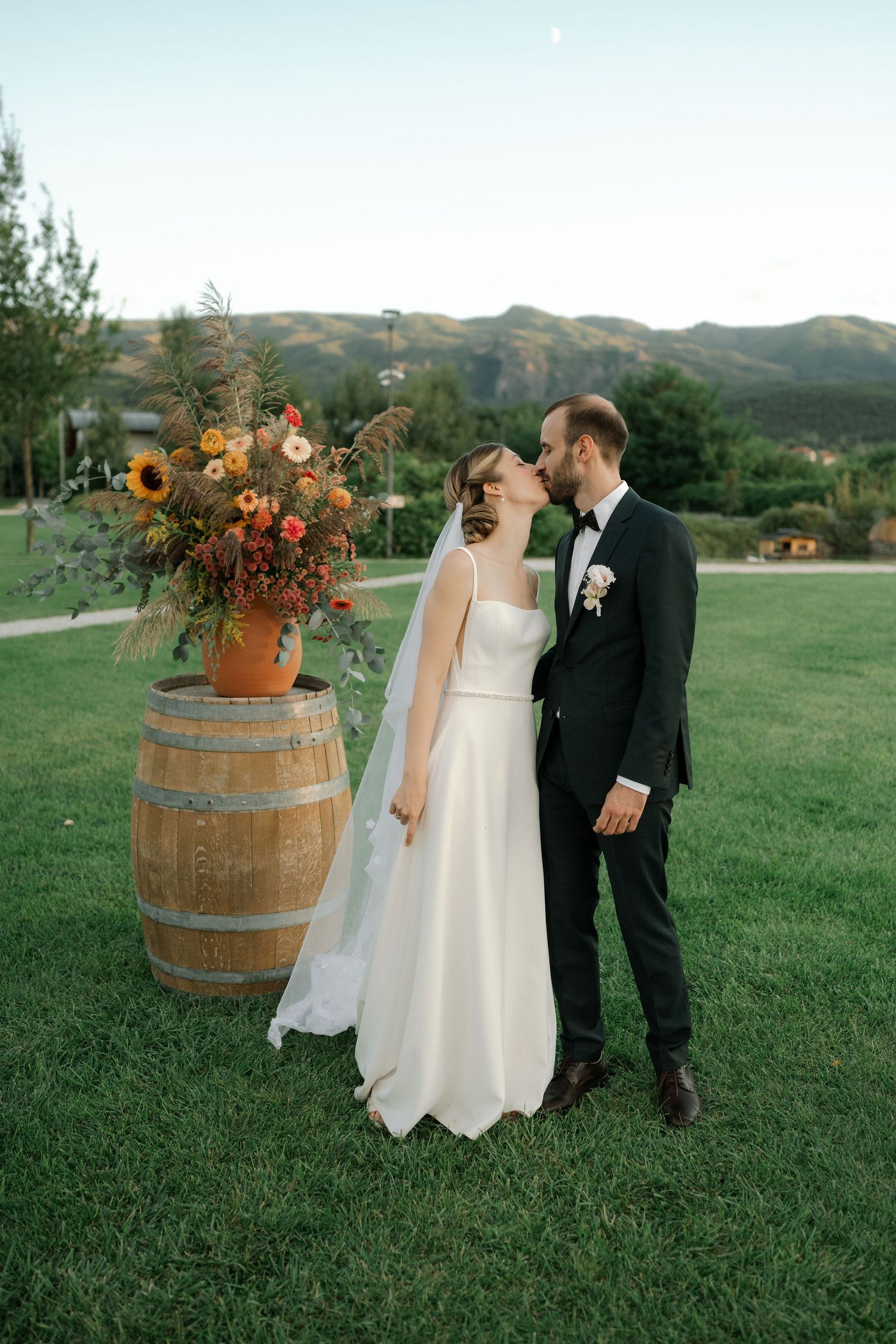 A romantic autumn wedding scene on a grassy hill with rustic wine barrels and orange floral arrangements at sunset.