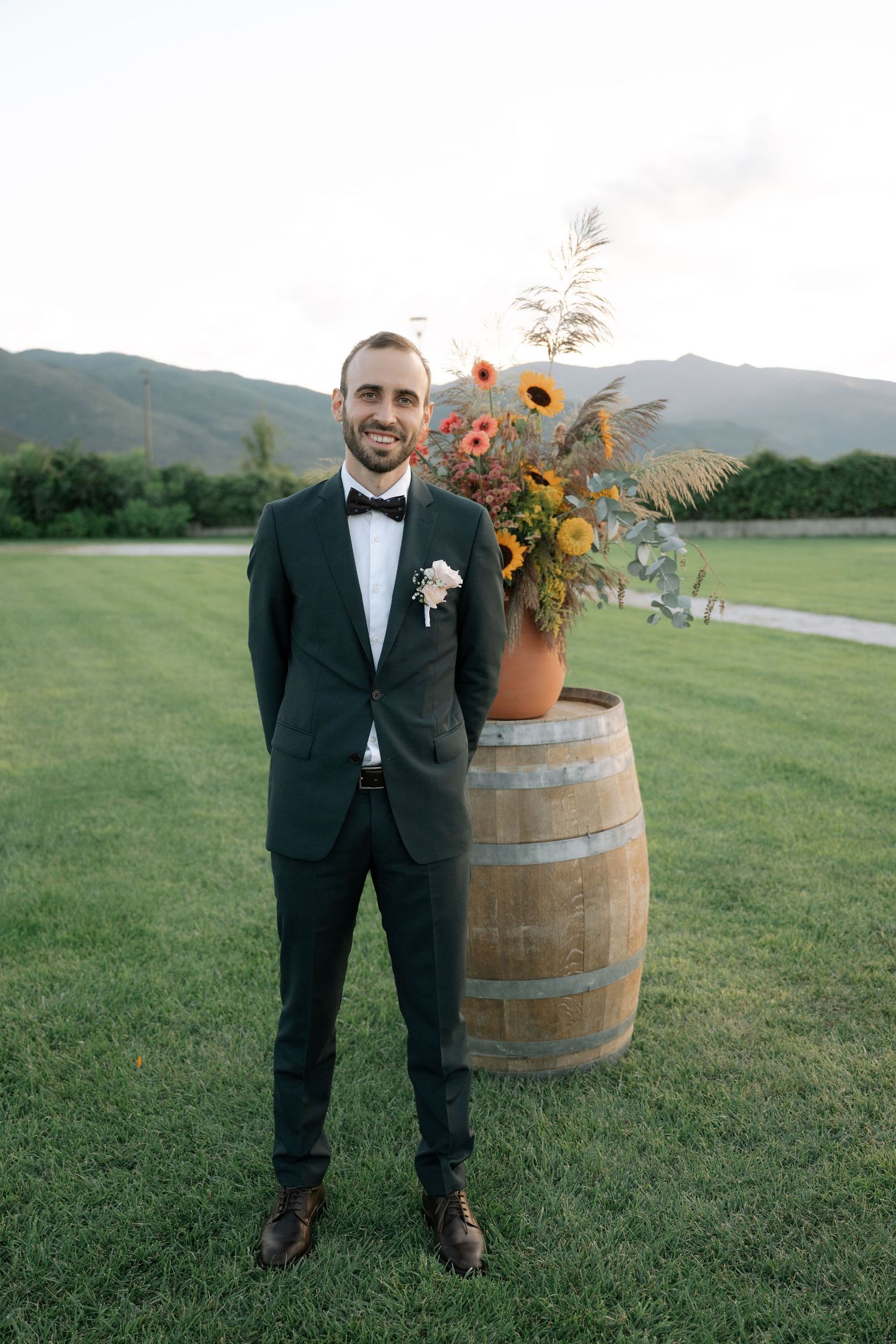 Wedding guest in dark tuxedo and bowtie stands beside rustic wine barrel with floral arrangement on outdoor lawn.