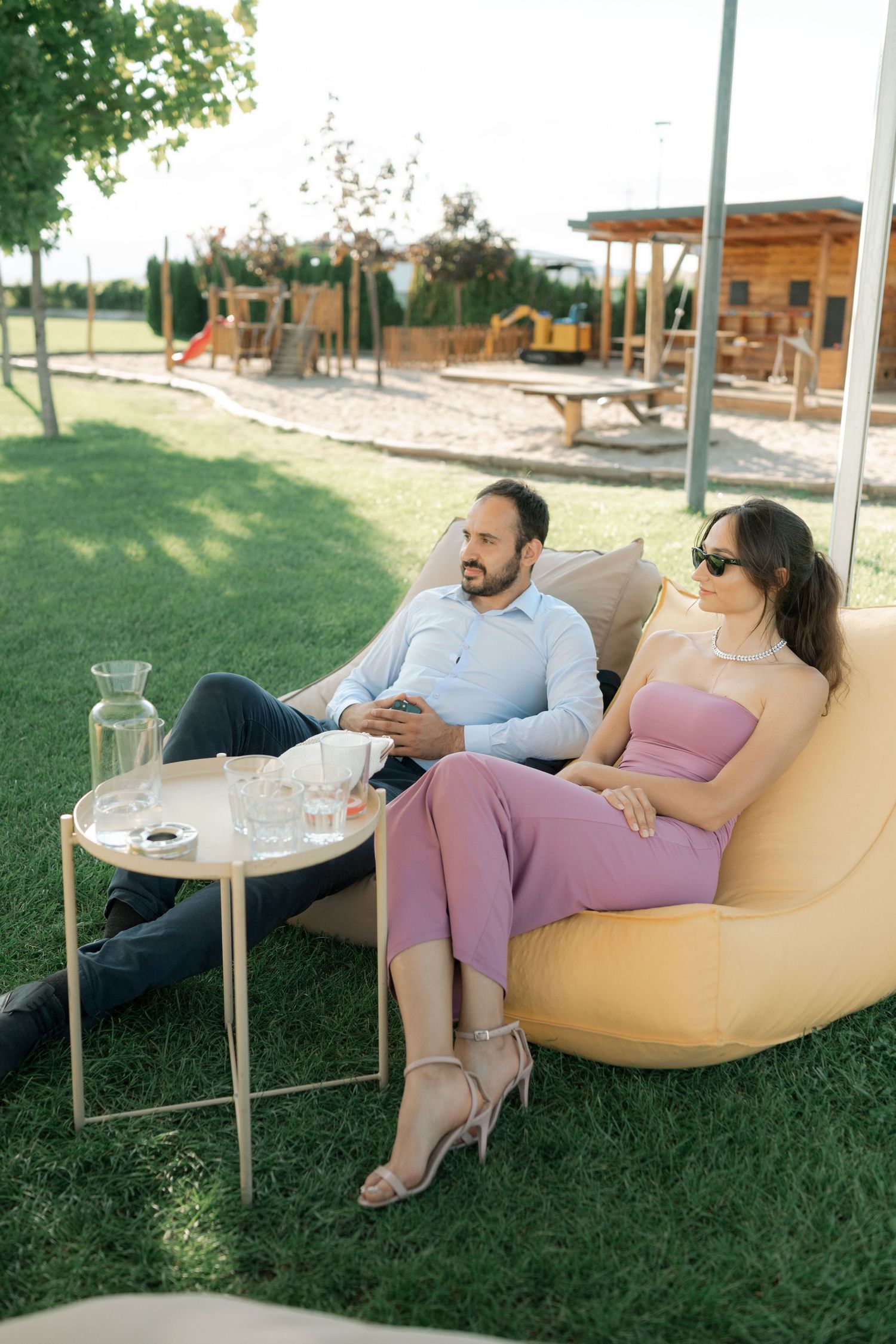 A couple relaxes together on a yellow outdoor lounge chair while looking at their phones in a sunny garden setting.