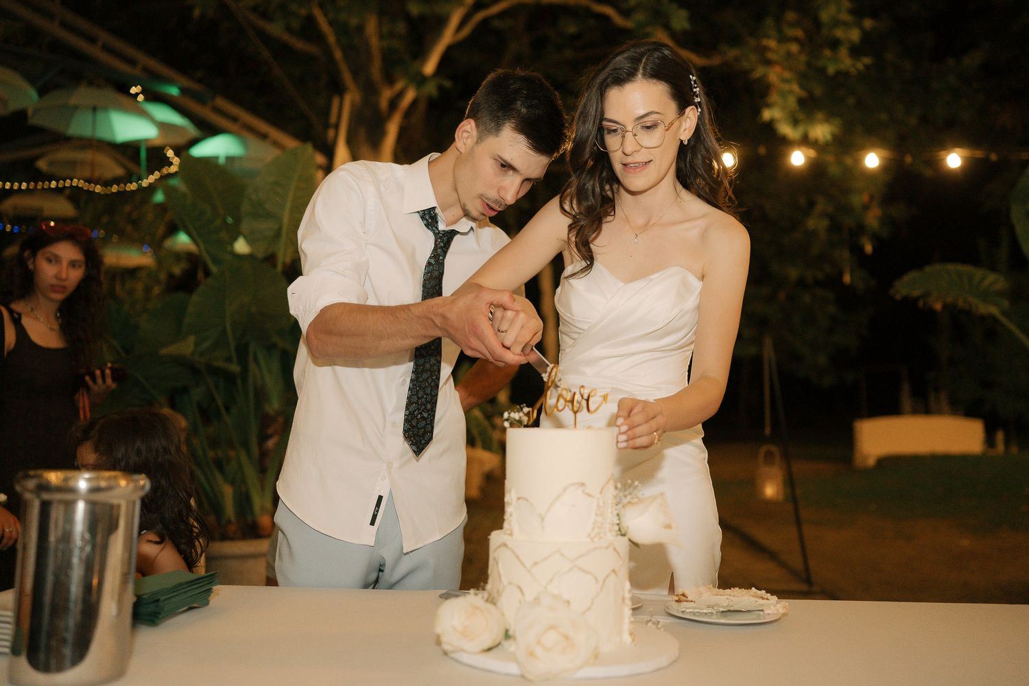 A couple shares a joyful moment while cutting their wedding cake at a nighttime outdoor reception.