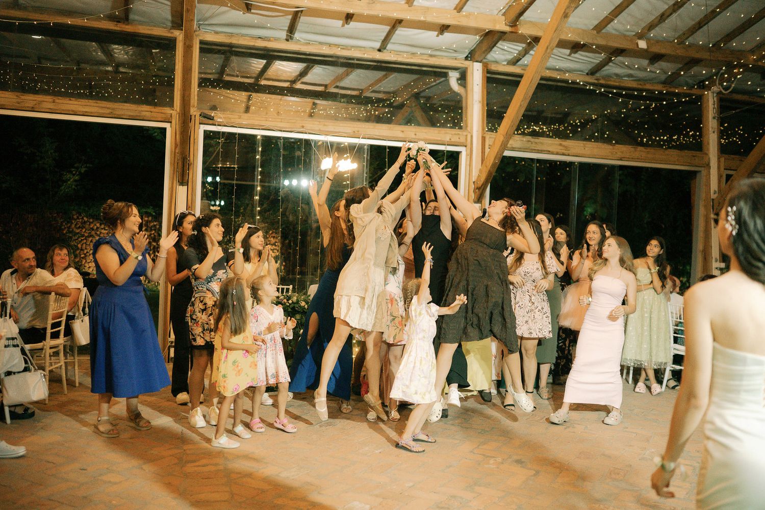 Wedding reception guests gather under wooden beams to catch the bouquet toss at a rustic barn venue.